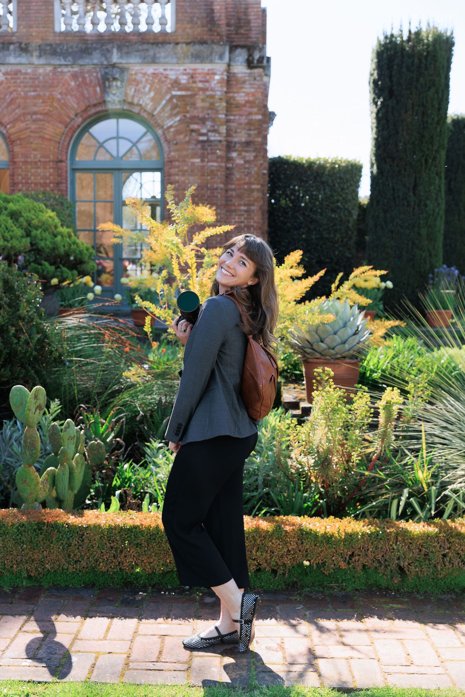 Well-dressed figure strikes playful pose in front of brick mansion with lush garden featuring cactus and cypress trees.