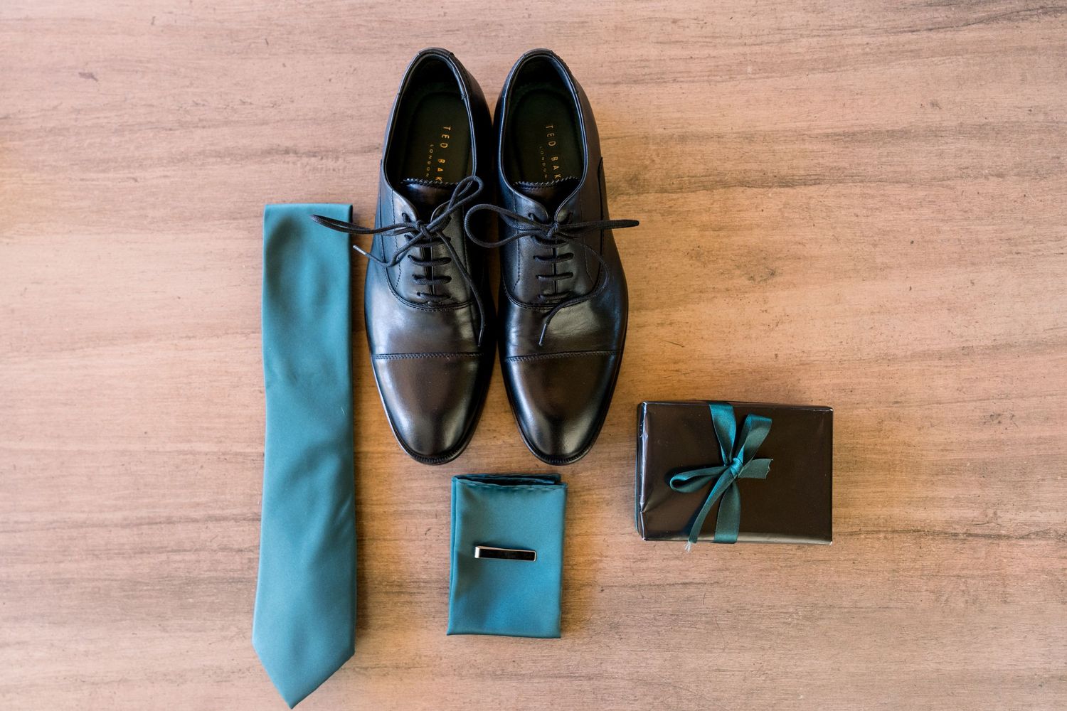Elegant groom accessories including black dress shoes, teal necktie, and small gift box arranged on wooden floor.