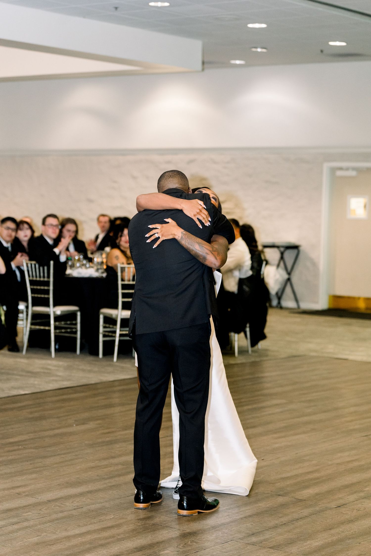A heartfelt embrace on the dance floor at an elegant wedding reception with guests seated at tables in the background.