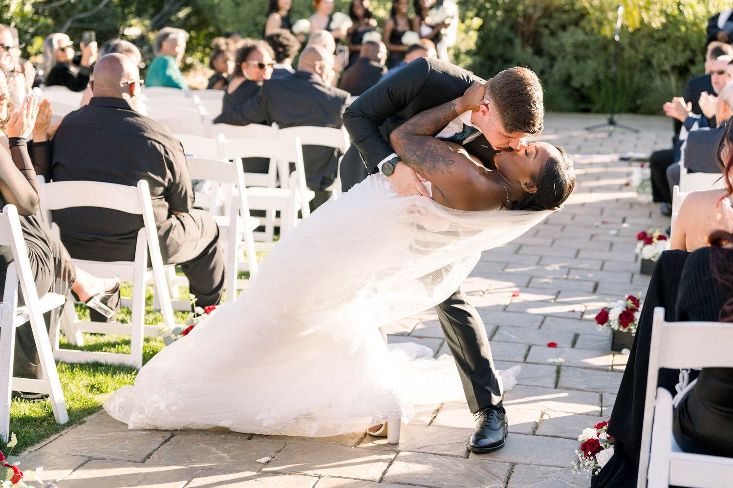 Romantic dip kiss during an outdoor wedding ceremony with guests seated in white chairs.