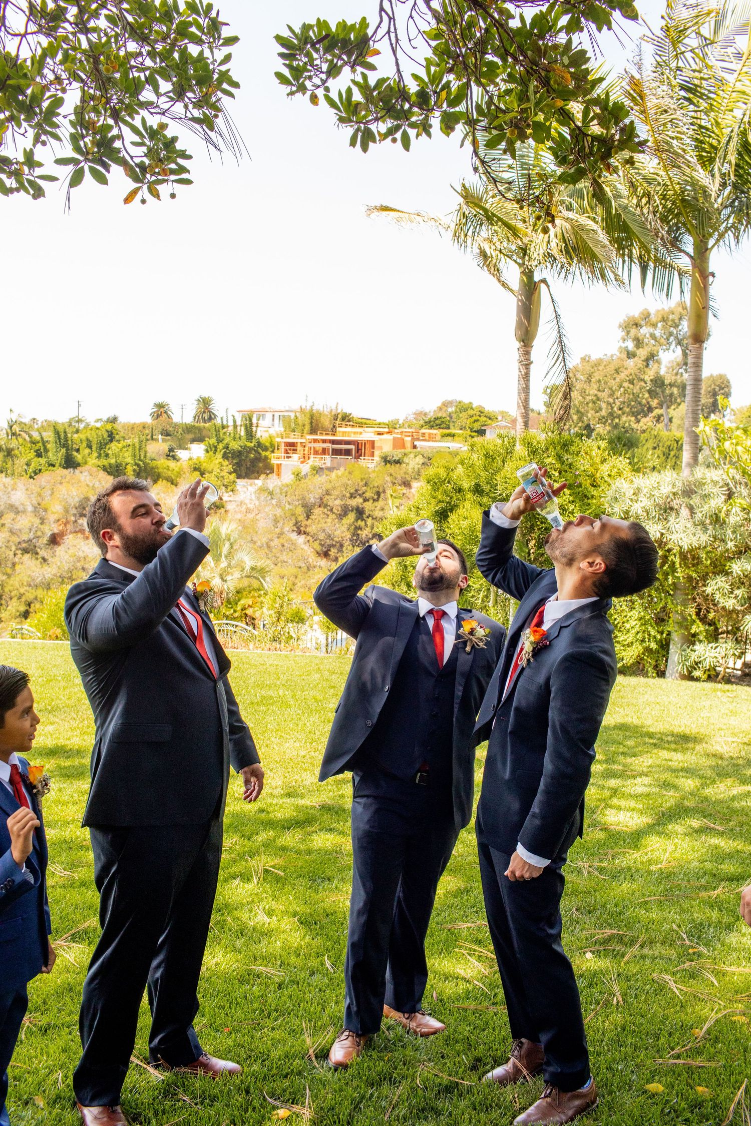 Groomsmen in navy suits taking celebratory shots together on a sunny lawn.