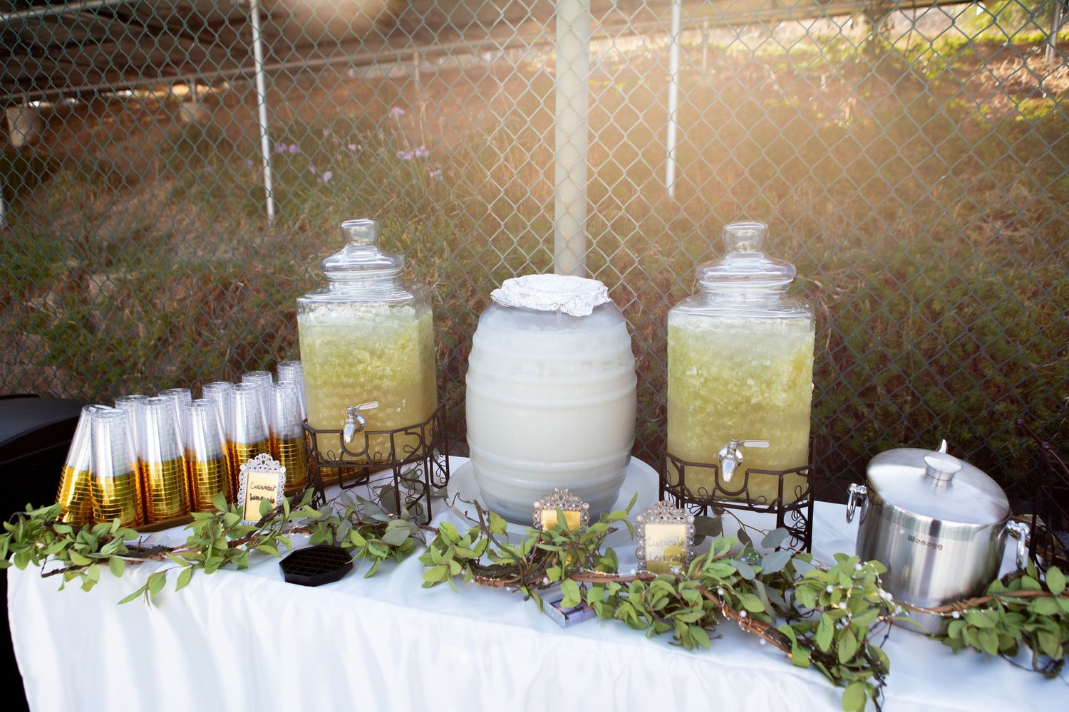 A beverage station with glass dispensers and decorative greenery set up for an outdoor wedding reception.