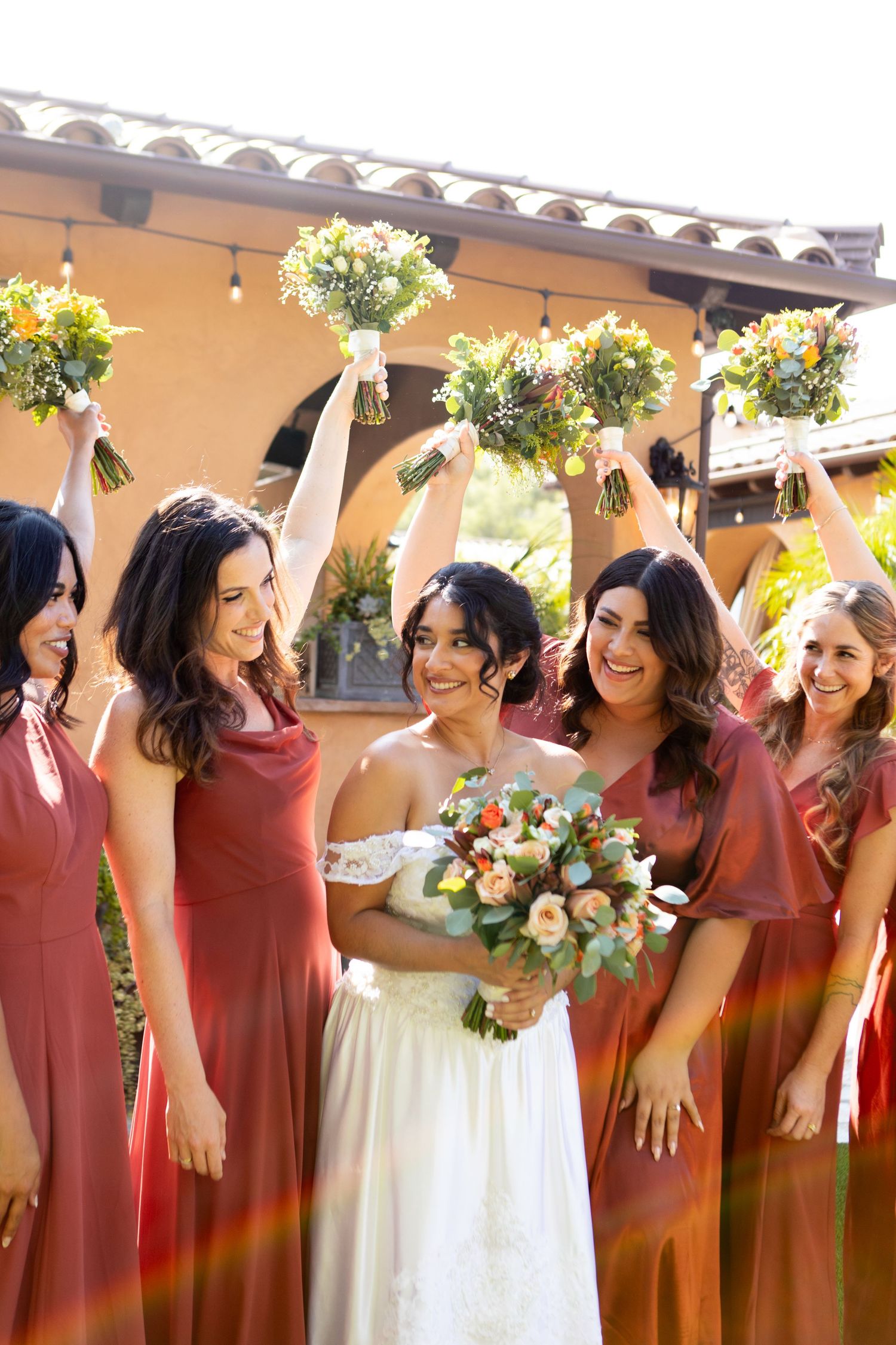 Wedding party celebrating with raised bouquets under Spanish-style architecture.