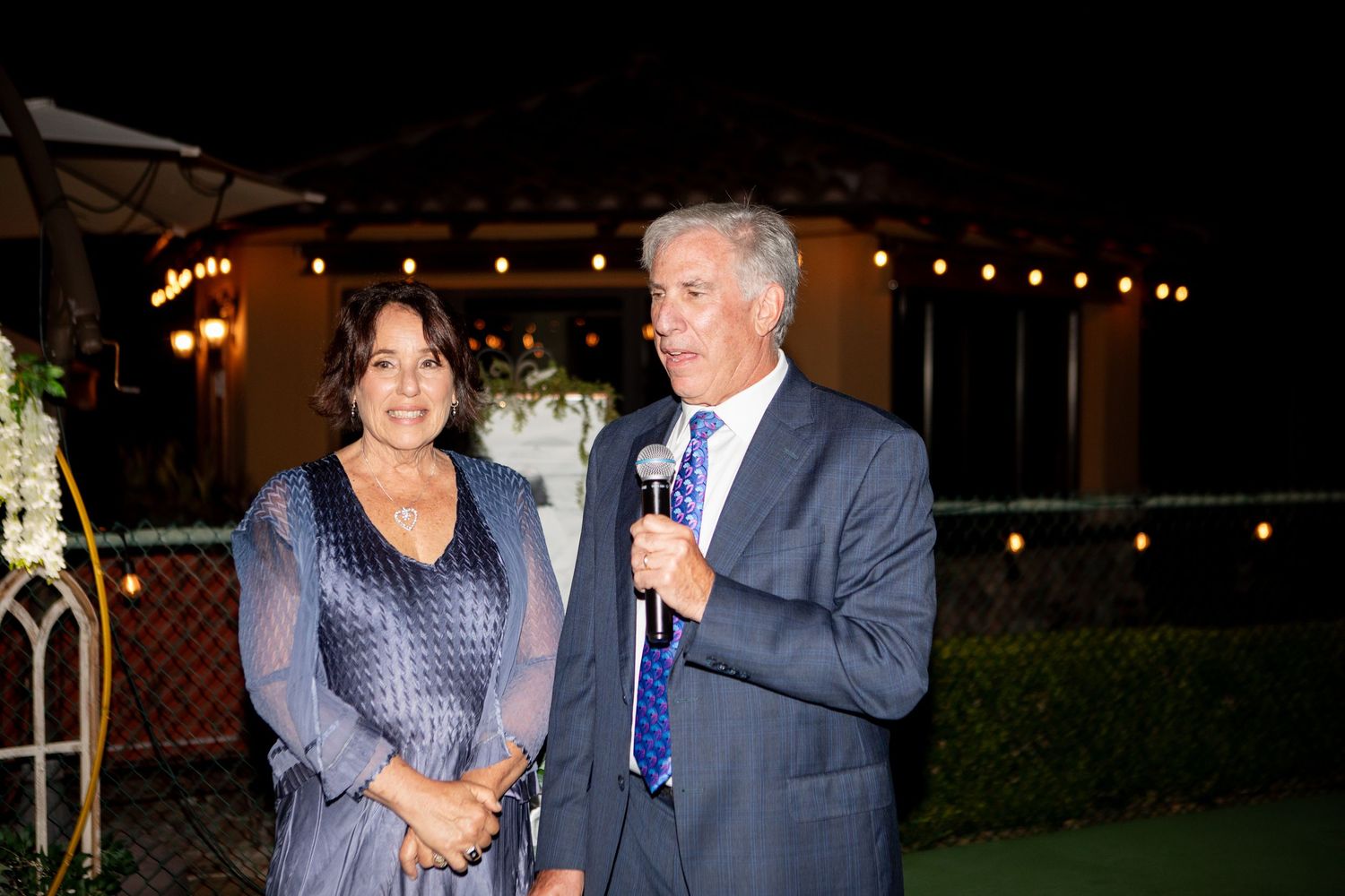 Two people speaking at an evening outdoor event with string lights and greenery in the background.