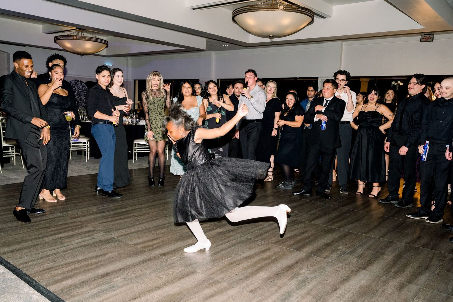 Person in black performing breakdance moves on wooden floor surrounded by wedding guests.