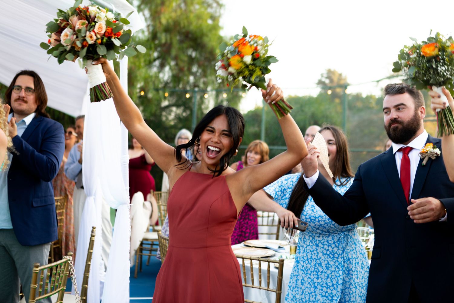 Wedding party celebrates with raised bouquets during outdoor ceremony with wooden chairs.