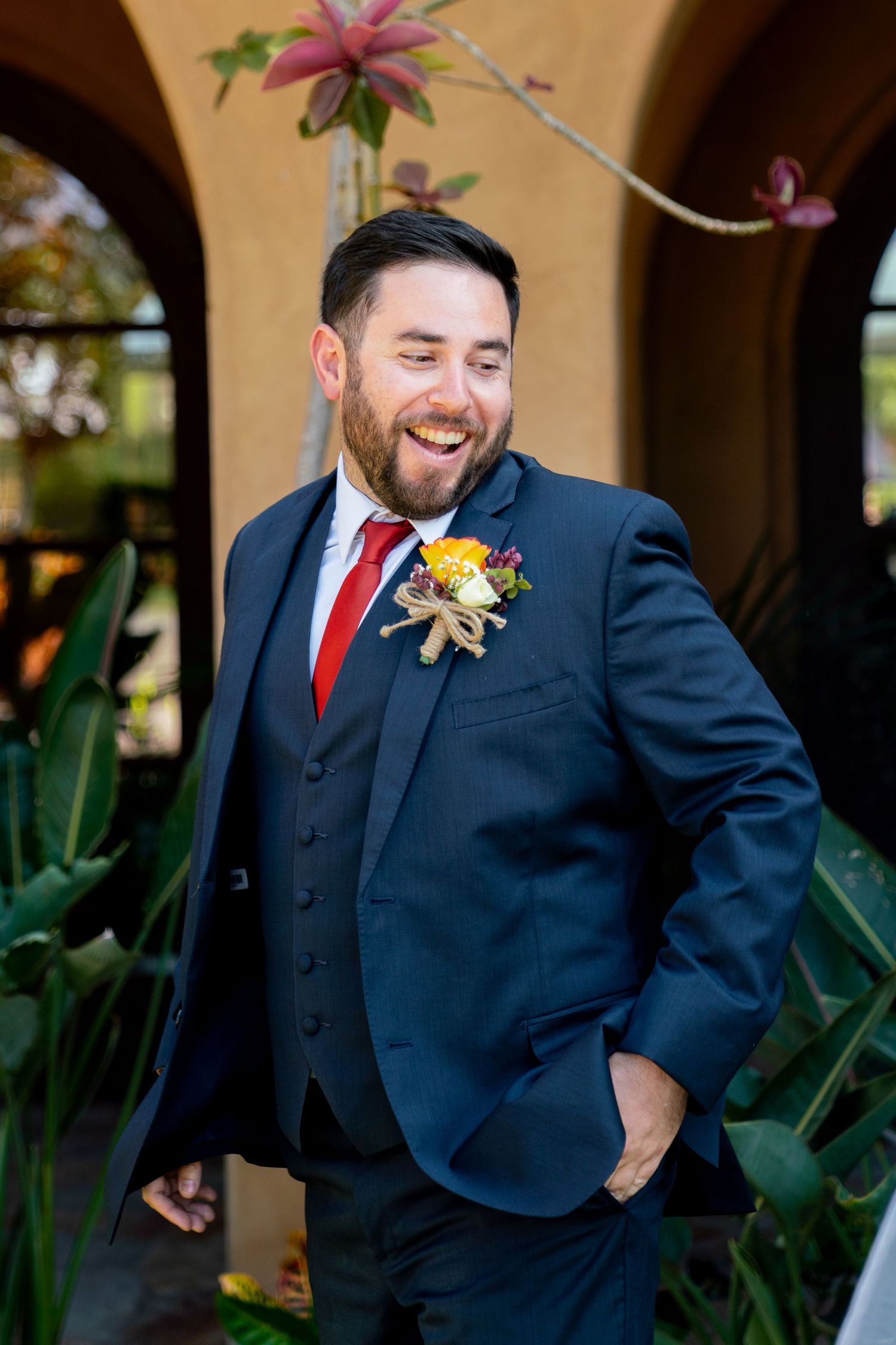Wedding attire featuring a navy suit with red tie and floral boutonniere against an archway.