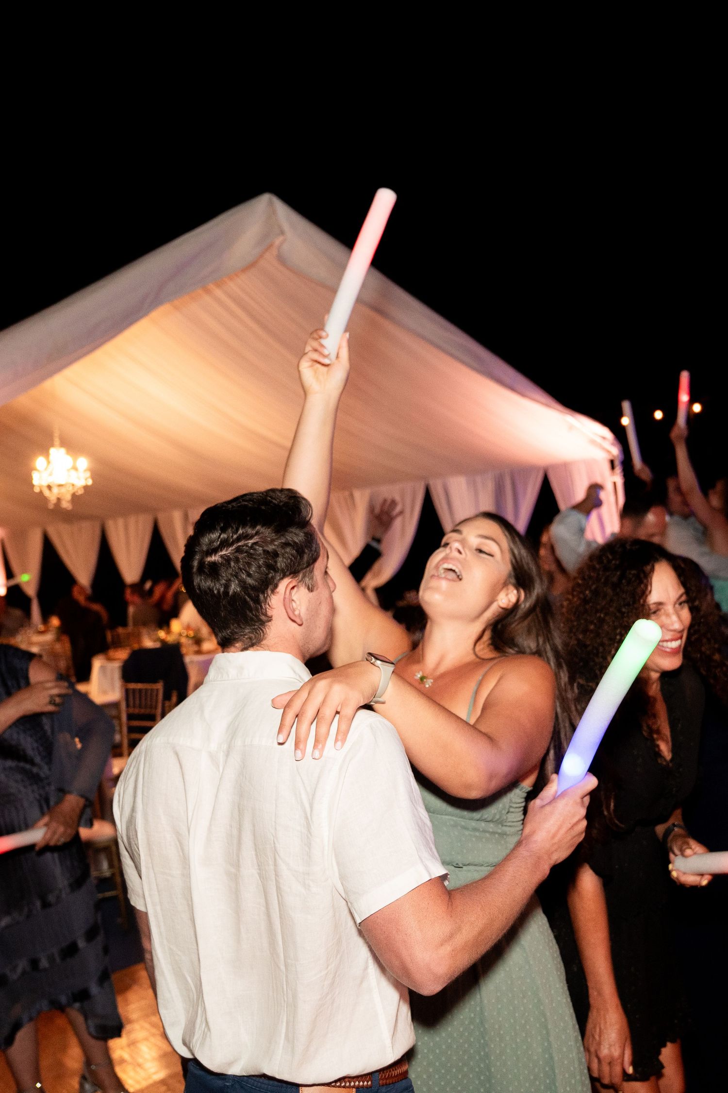 Guests dancing with glow sticks under white tent at nighttime wedding reception.