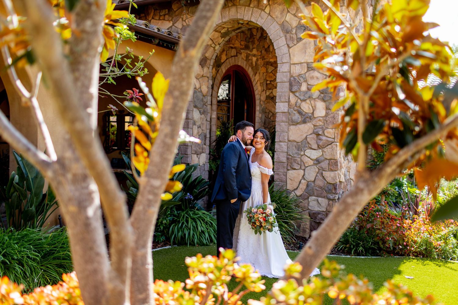 Newlyweds share a romantic moment by a stone archway surrounded by autumn foliage.