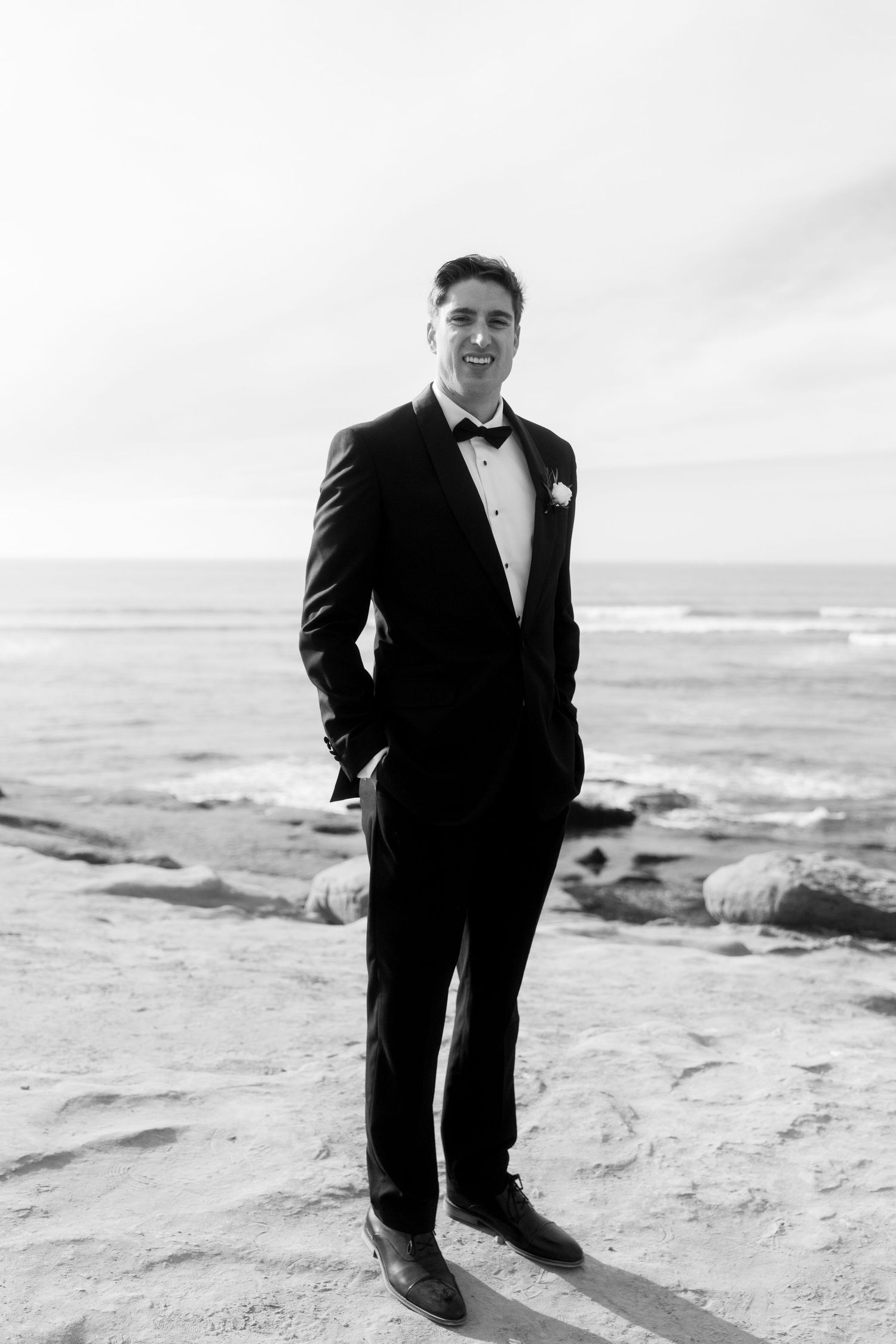A black and white portrait of a groom in a tuxedo standing on coastal rocks with ocean waves behind him.
