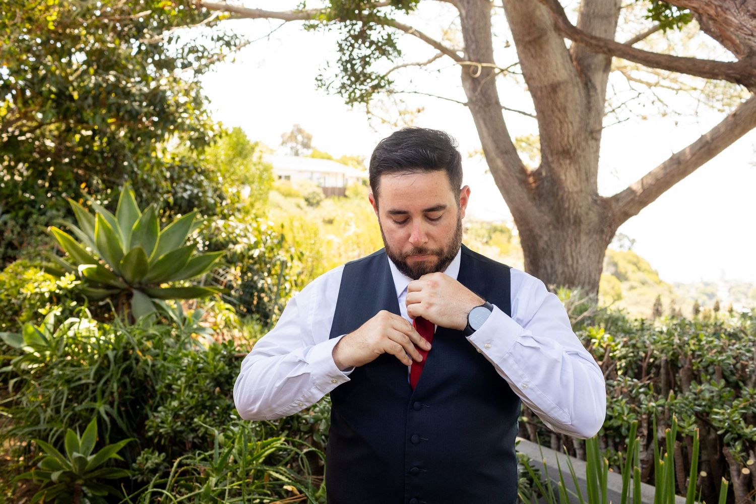 A person in business attire adjusts their tie while standing in a lush garden setting with trees and plants.