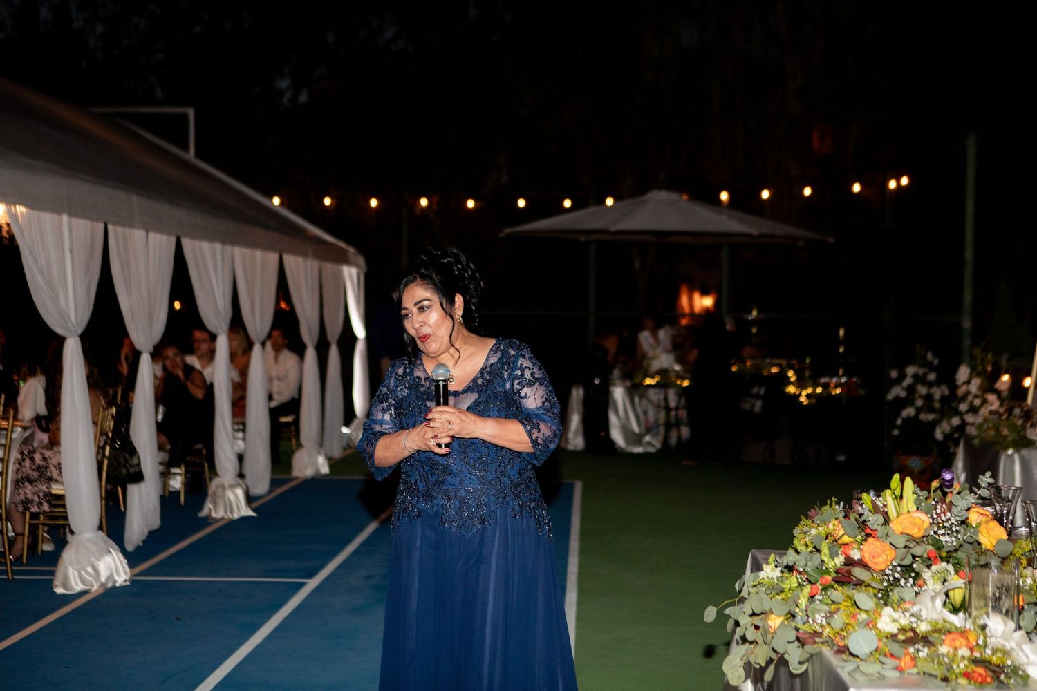 Guest in navy blue dress speaks at evening outdoor reception.
