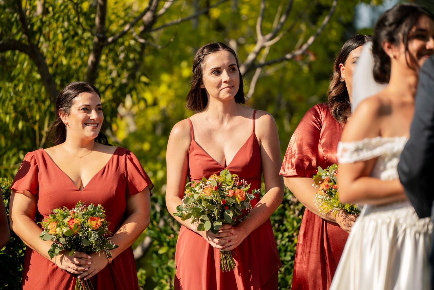 Bridesmaids in coral dresses hold yellow flower bouquets during an outdoor wedding ceremony.