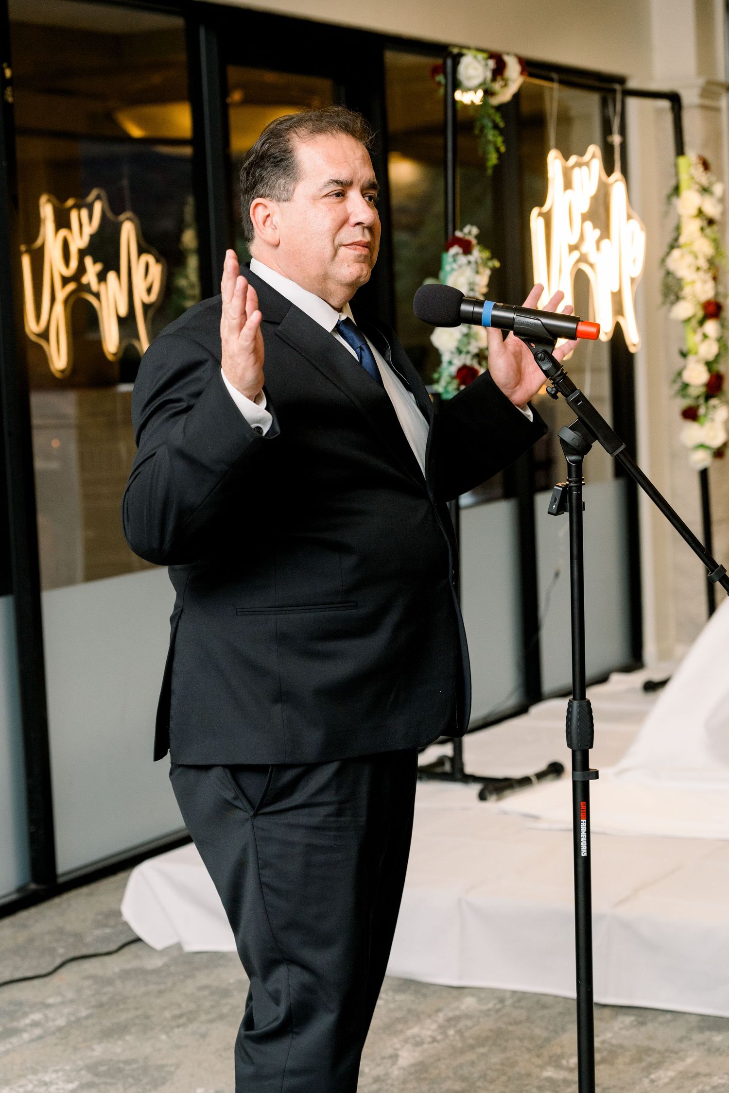 Speaker in black suit addresses wedding guests at evening reception with illuminated signage.