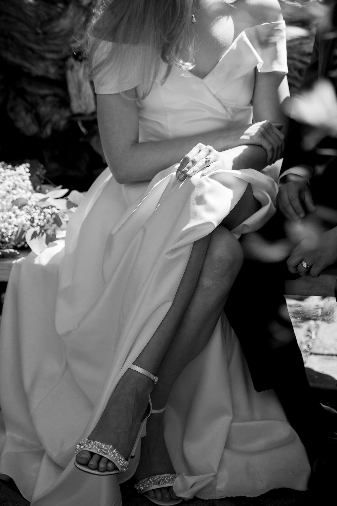 Black and white photo showing a bride in flowing white dress seated outdoors, with a decorated leg garter visible.