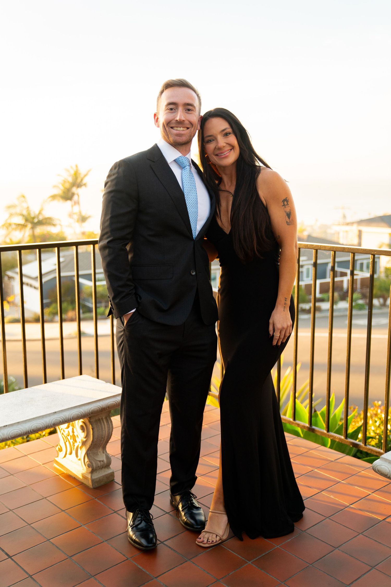 A couple in formal attire poses on a sunset terrace with palm trees in the background.
