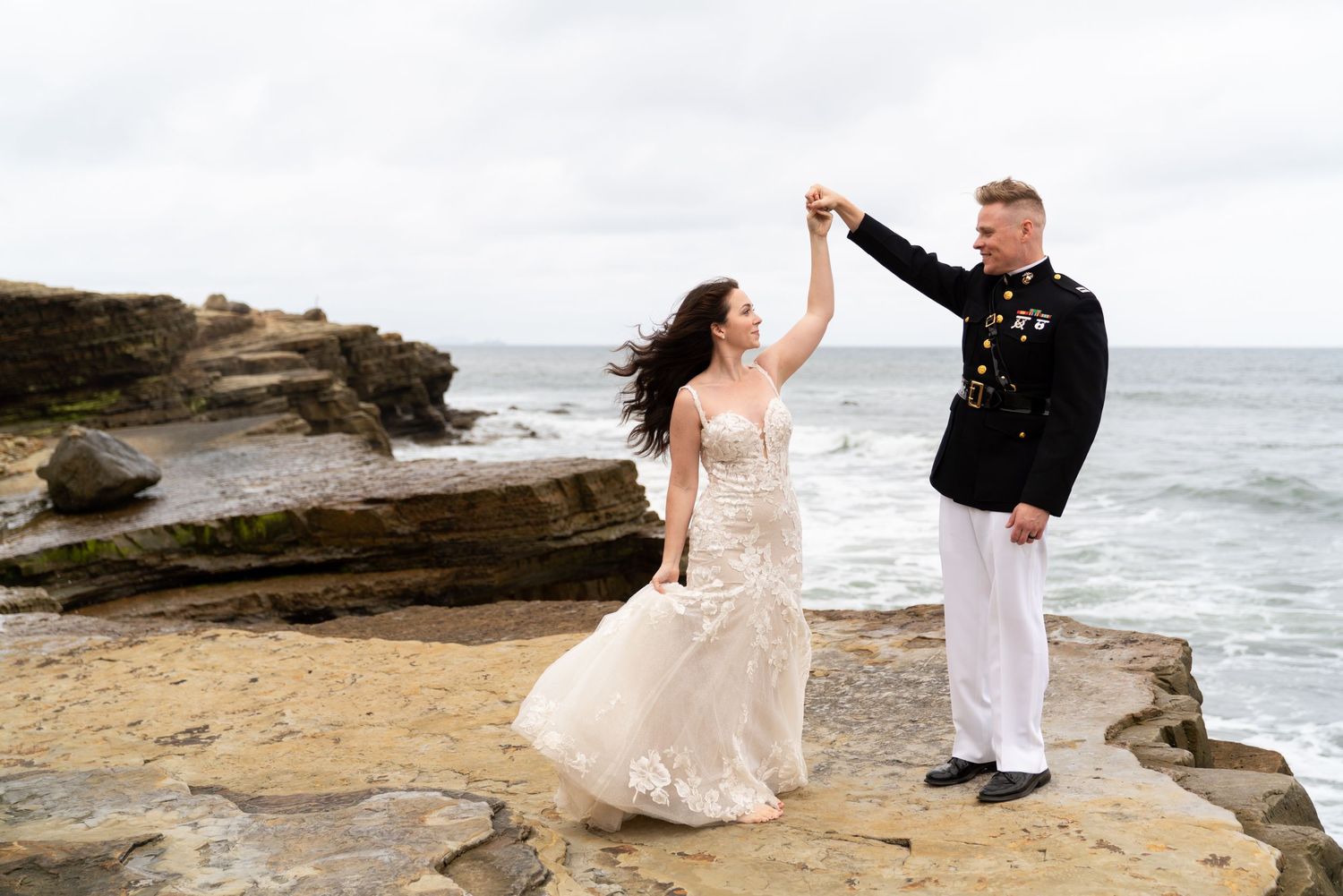 A romantic wedding couple dancing together on rocky ocean cliffs against a dramatic coastal backdrop.