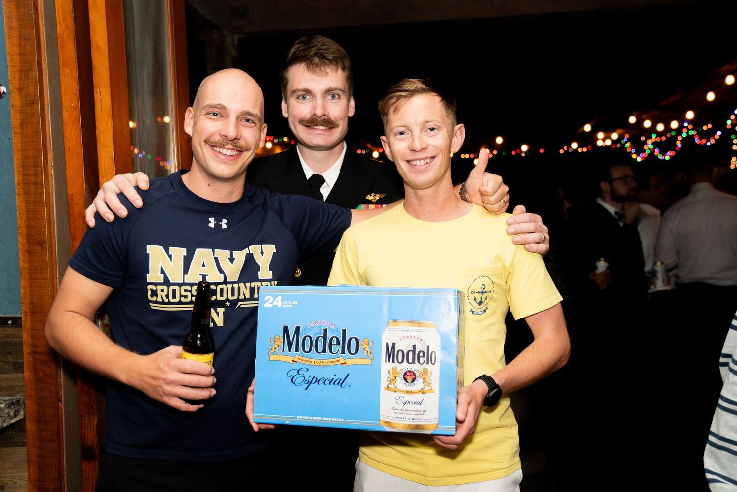 Three friends pose with Modelo beer box at casual gathering.