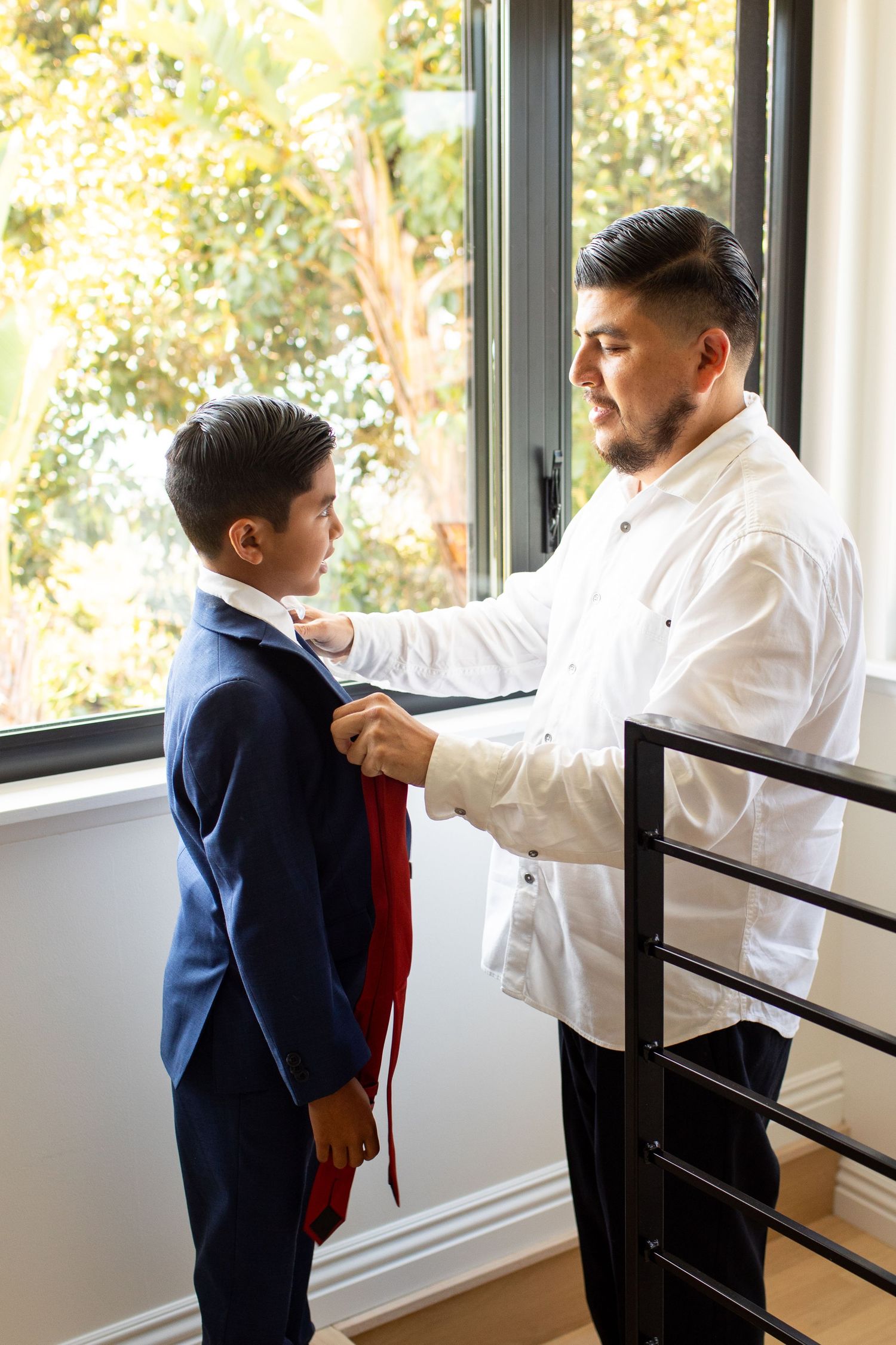 A tender moment as someone helps adjust a young boy's navy suit tie near a bright window.