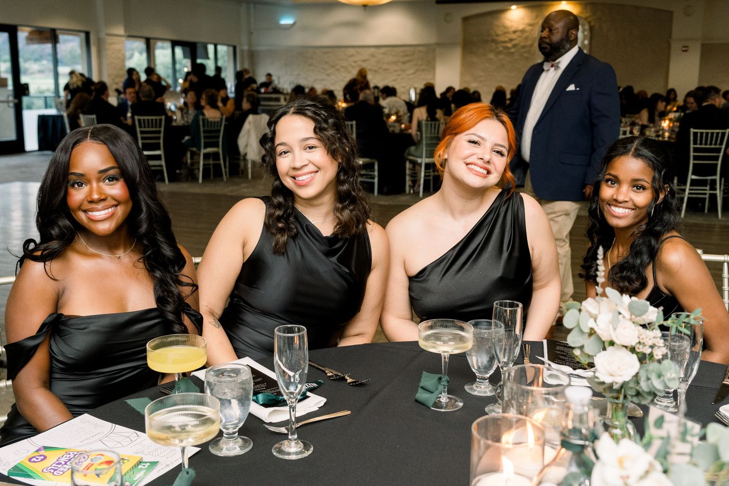 Wedding reception guests in black dresses smiling at a formal dinner table.