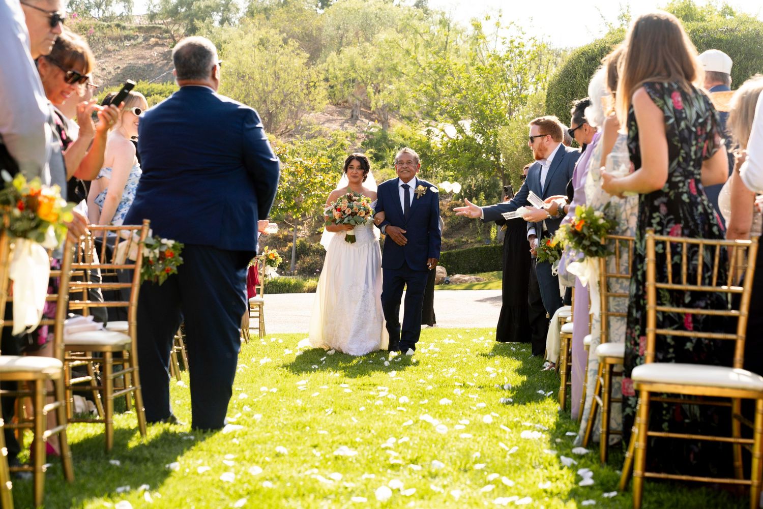 An outdoor wedding ceremony takes place on a sunny lawn with gold chairs and floral decorations.