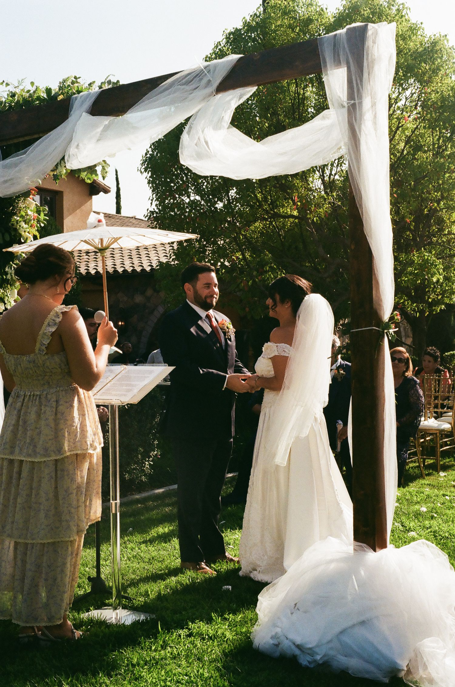 An outdoor wedding ceremony under a flowing white fabric canopy in a garden setting.