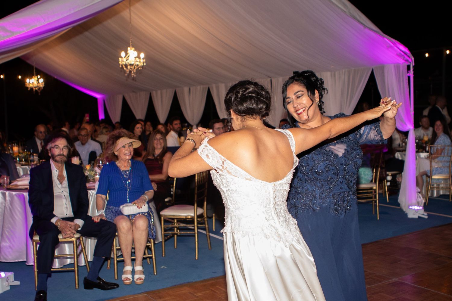 Reception dance moment captured under purple-lit tent with white draping and chandelier.
