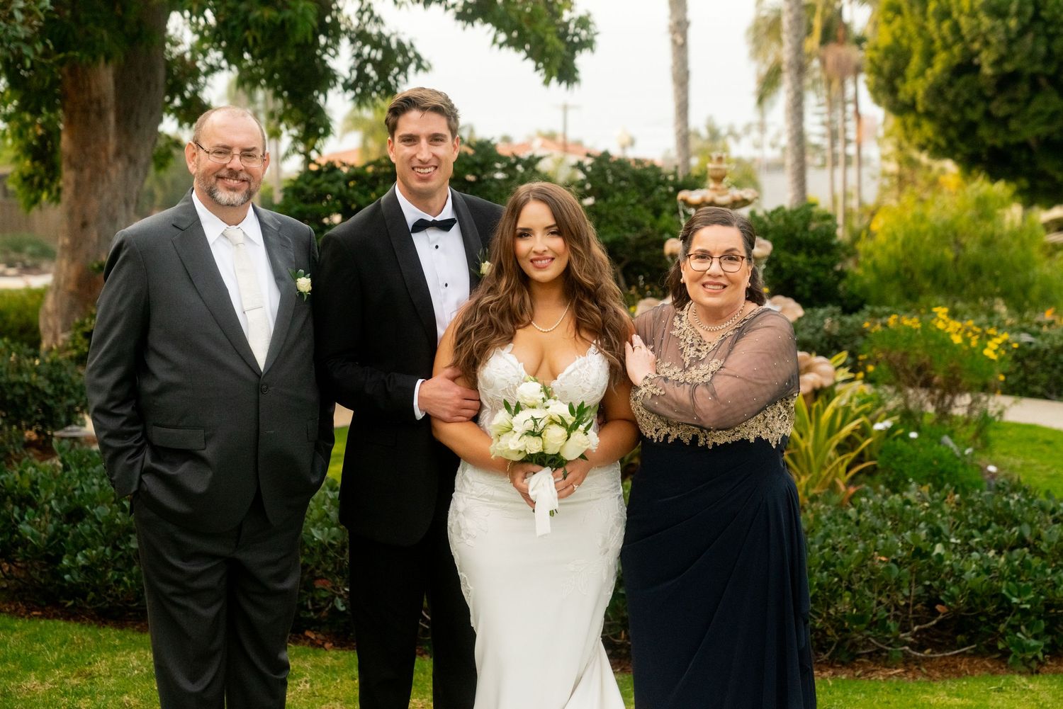 Wedding portrait of couple with family members in formal attire against garden background with palm trees.