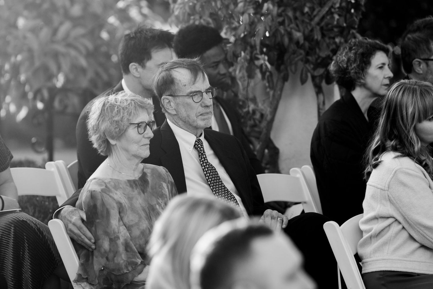 Guests seated outdoors at a formal gathering watch attentively while holding programs in their hands.