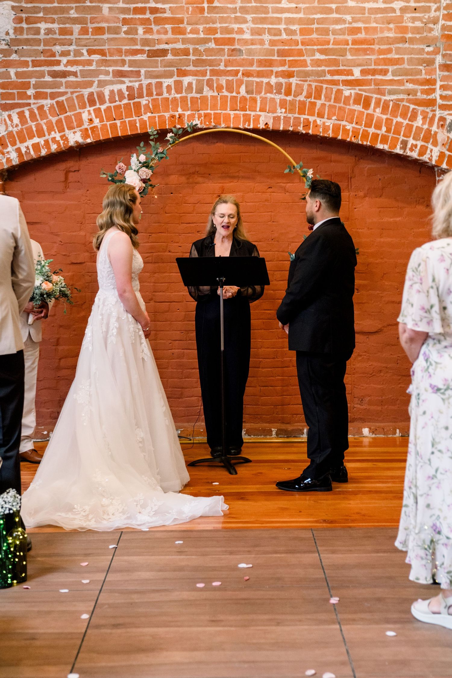 Wedding ceremony takes place under a decorative arch against exposed brick wall in an indoor venue.