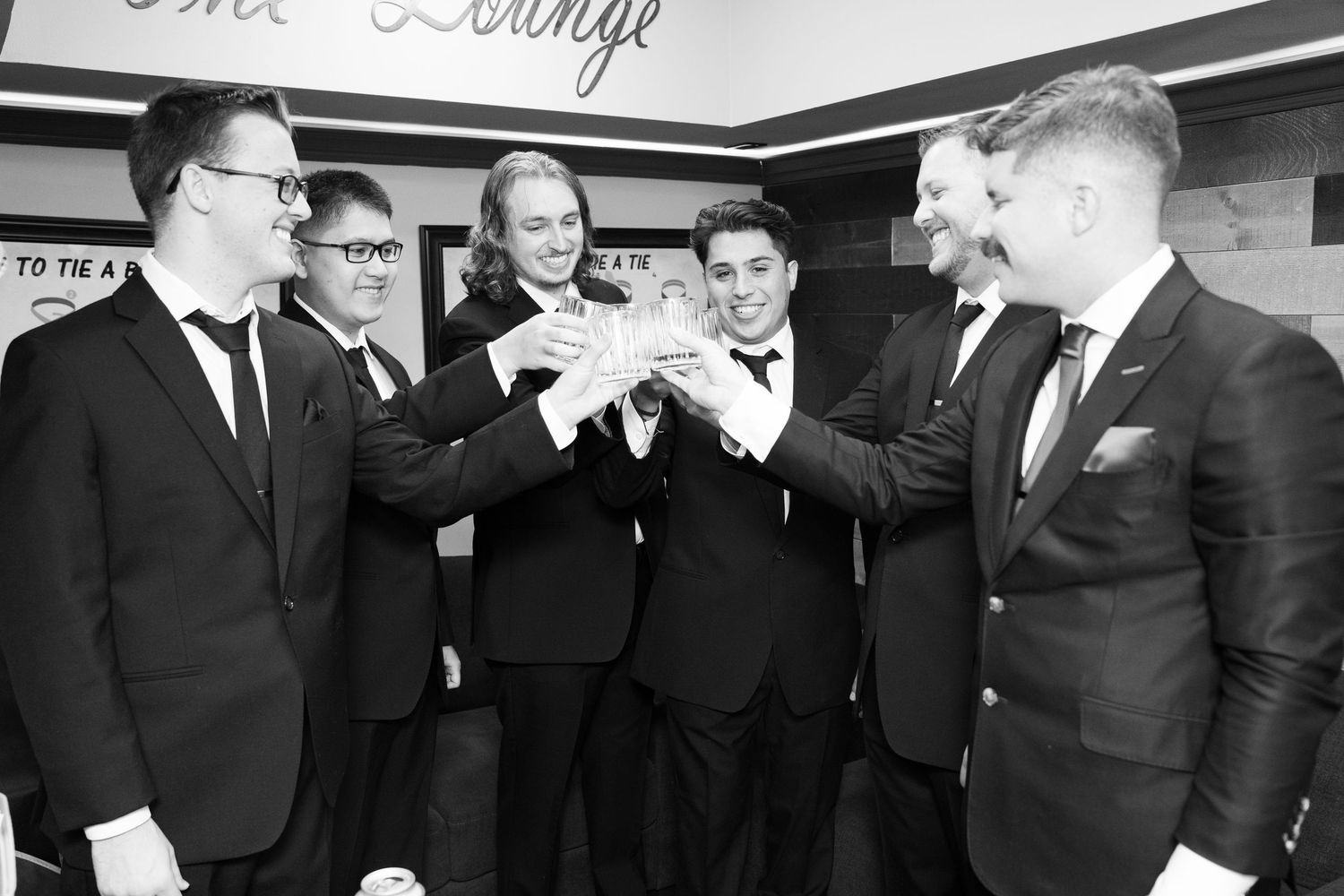 Groomsmen in black suits toasting with drinks during pre-wedding celebration.