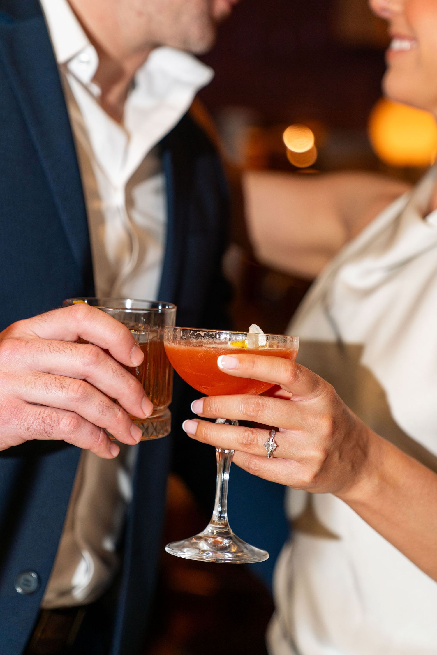 Two hands clinking cocktail glasses together in a cozy bar setting, with one person wearing a blue blazer.