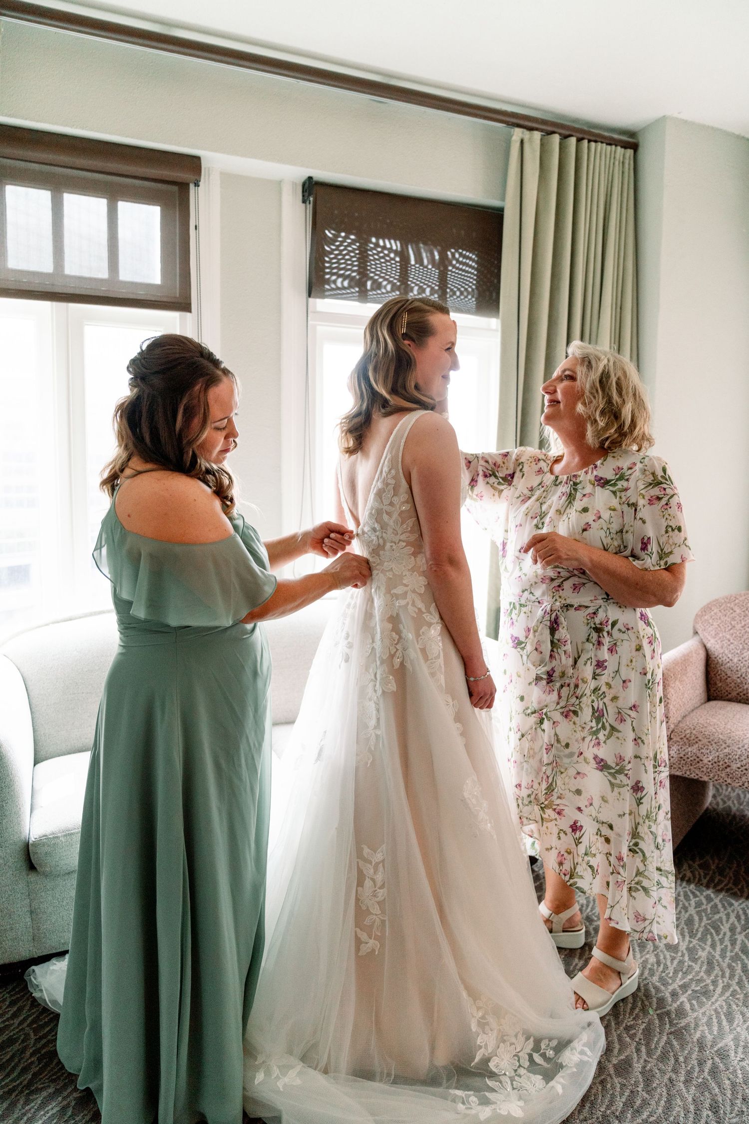 A bridal party helps with final preparations in a room with soft natural light and flowing curtains.