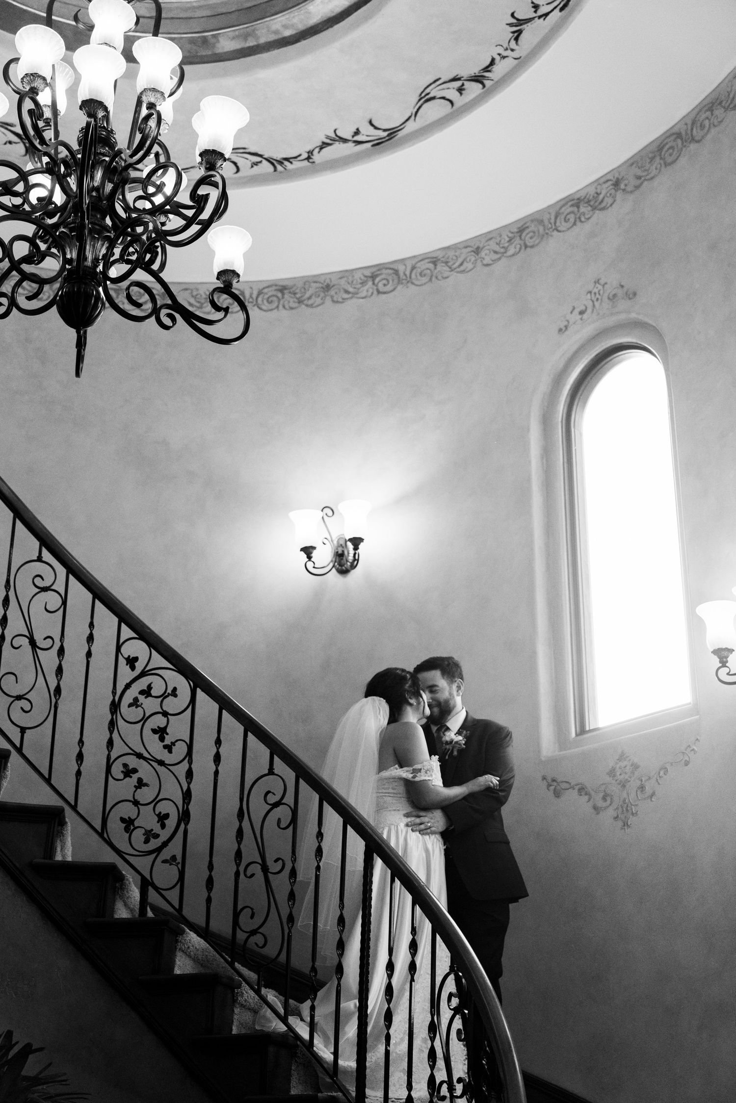 Black and white photo of couple on elegant spiral staircase with ornate chandelier and iron railing.