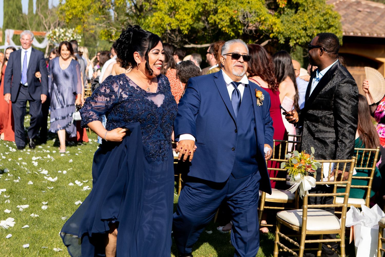 Wedding party members in navy blue attire walk across a lawn scattered with white petals.