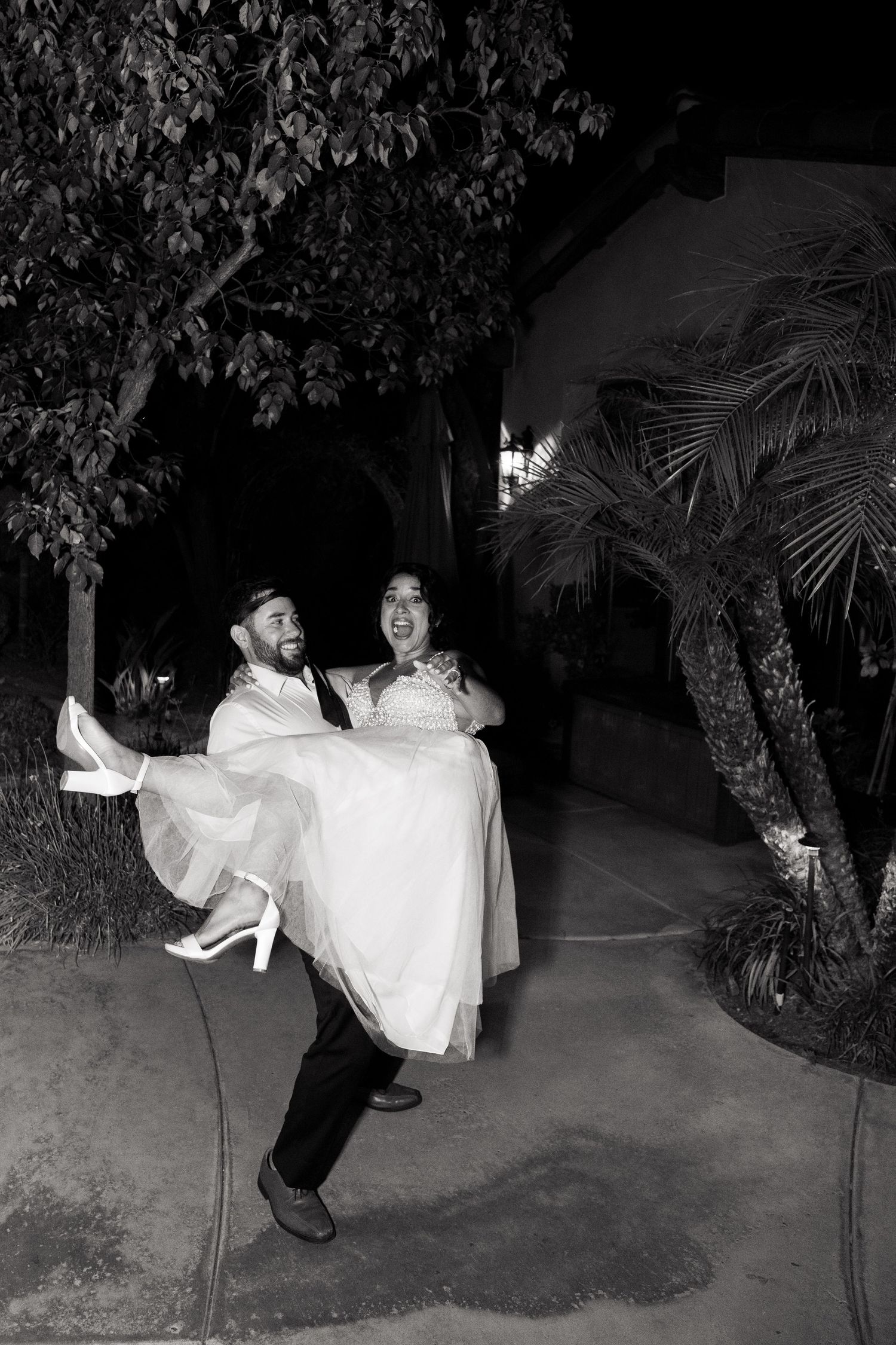 Black and white photo of couple sharing playful moment as groom lifts bride on outdoor walkway.