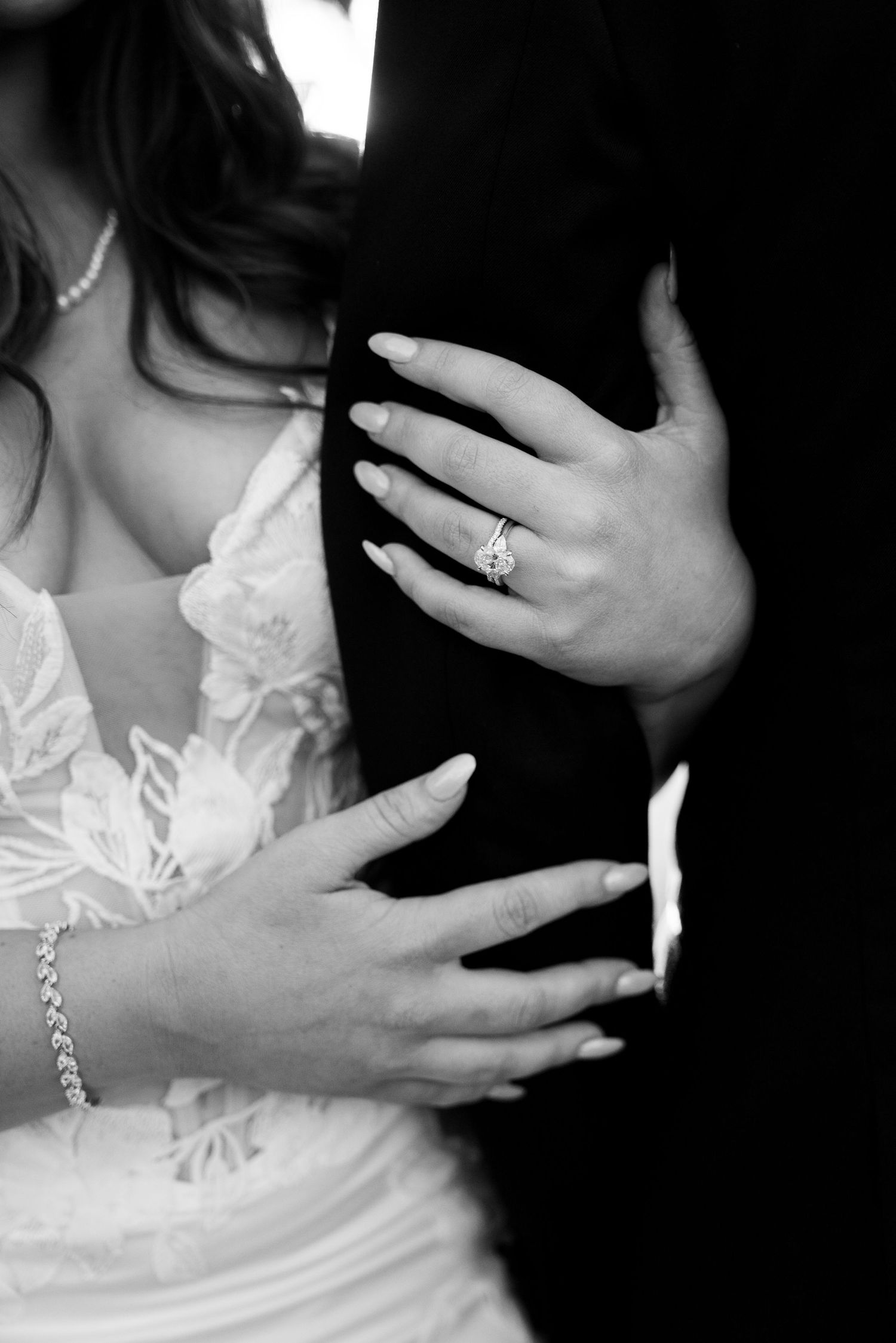 Close-up black and white shot showcases bride's hands with engagement ring resting on groom's suit.