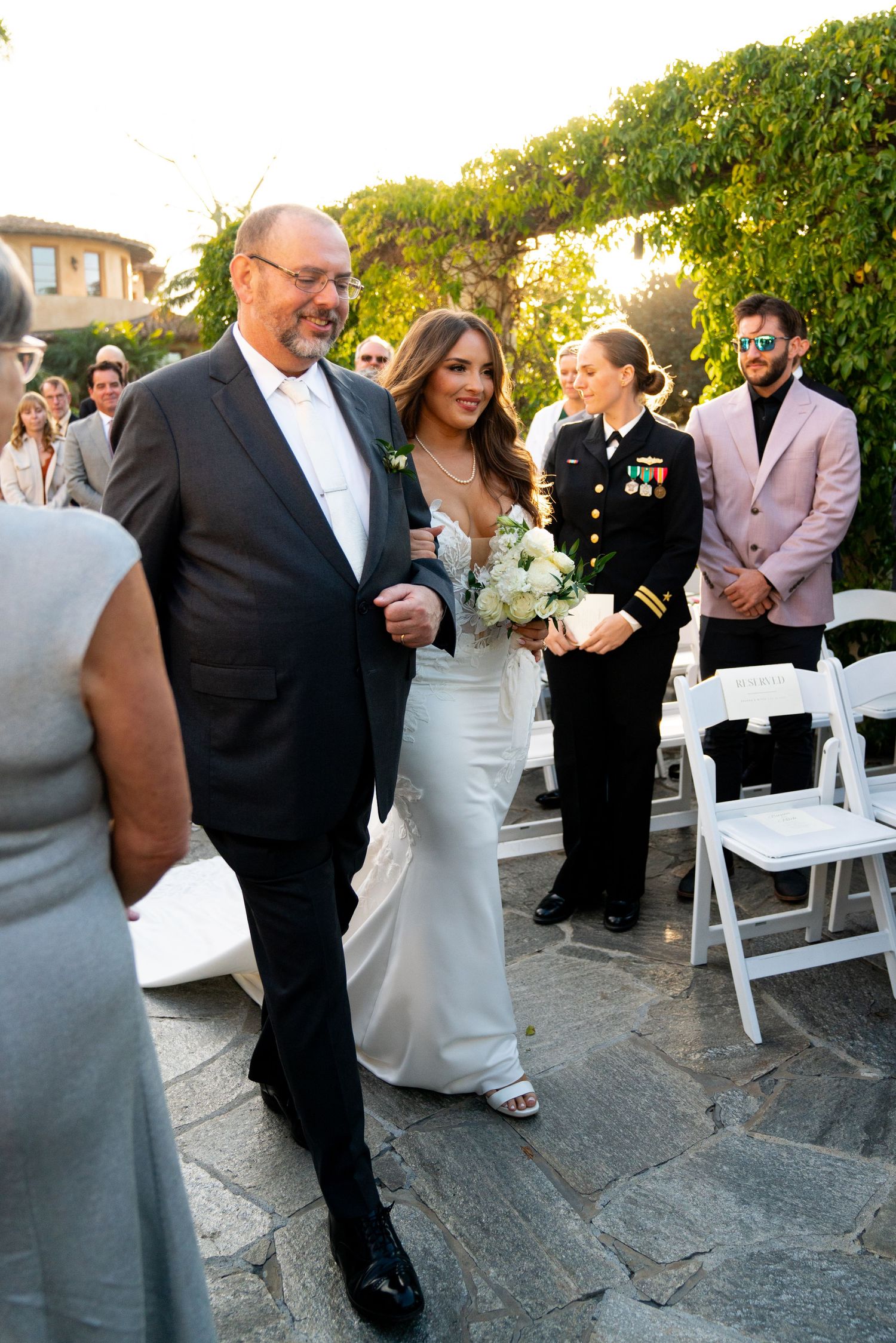 Wedding processional with couple walking down stone pathway surrounded by guests and white folding chairs.