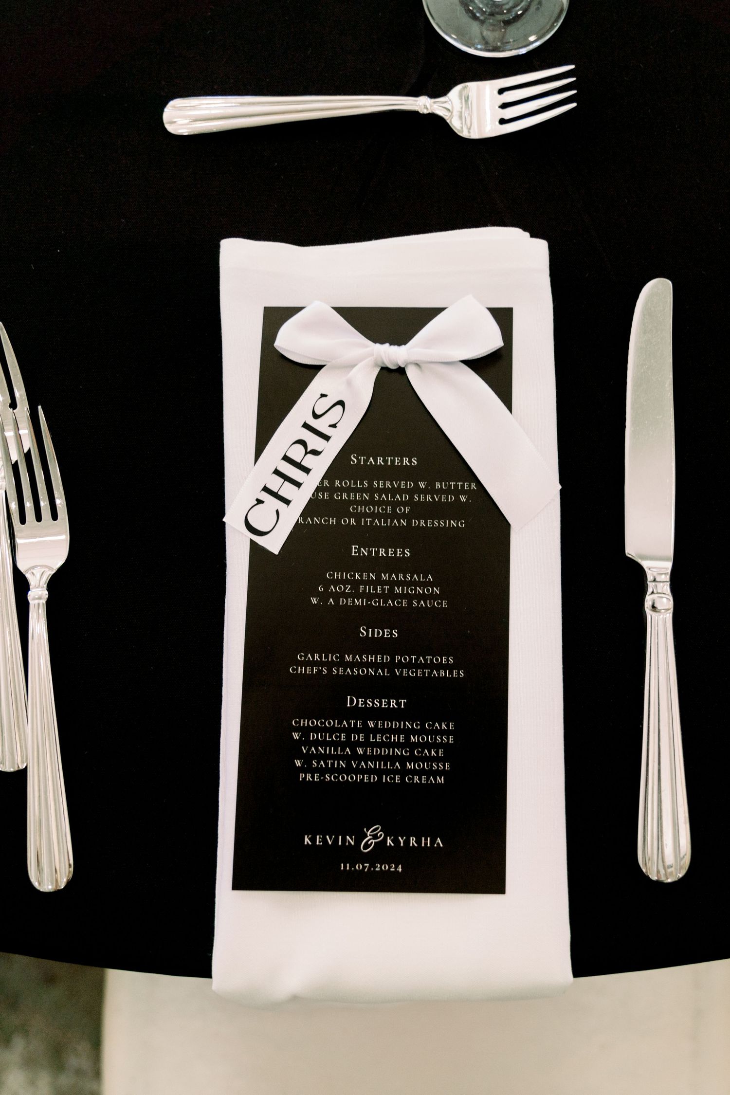 Wedding menu card with white bow ribbon displayed on black tablecloth with silverware.