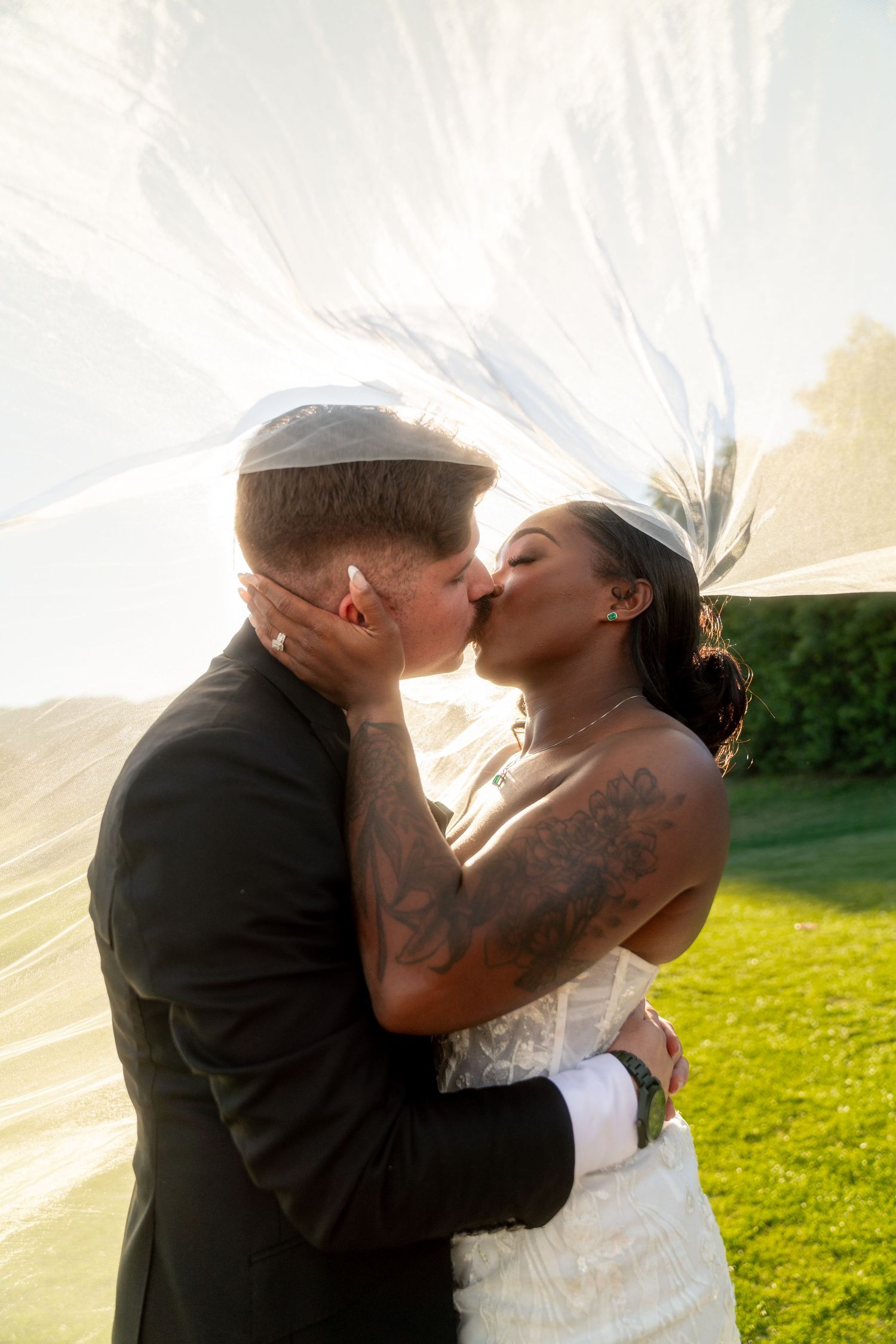 Intimate wedding moment captured under flowing veil with sunlight creating a dreamy backdrop.