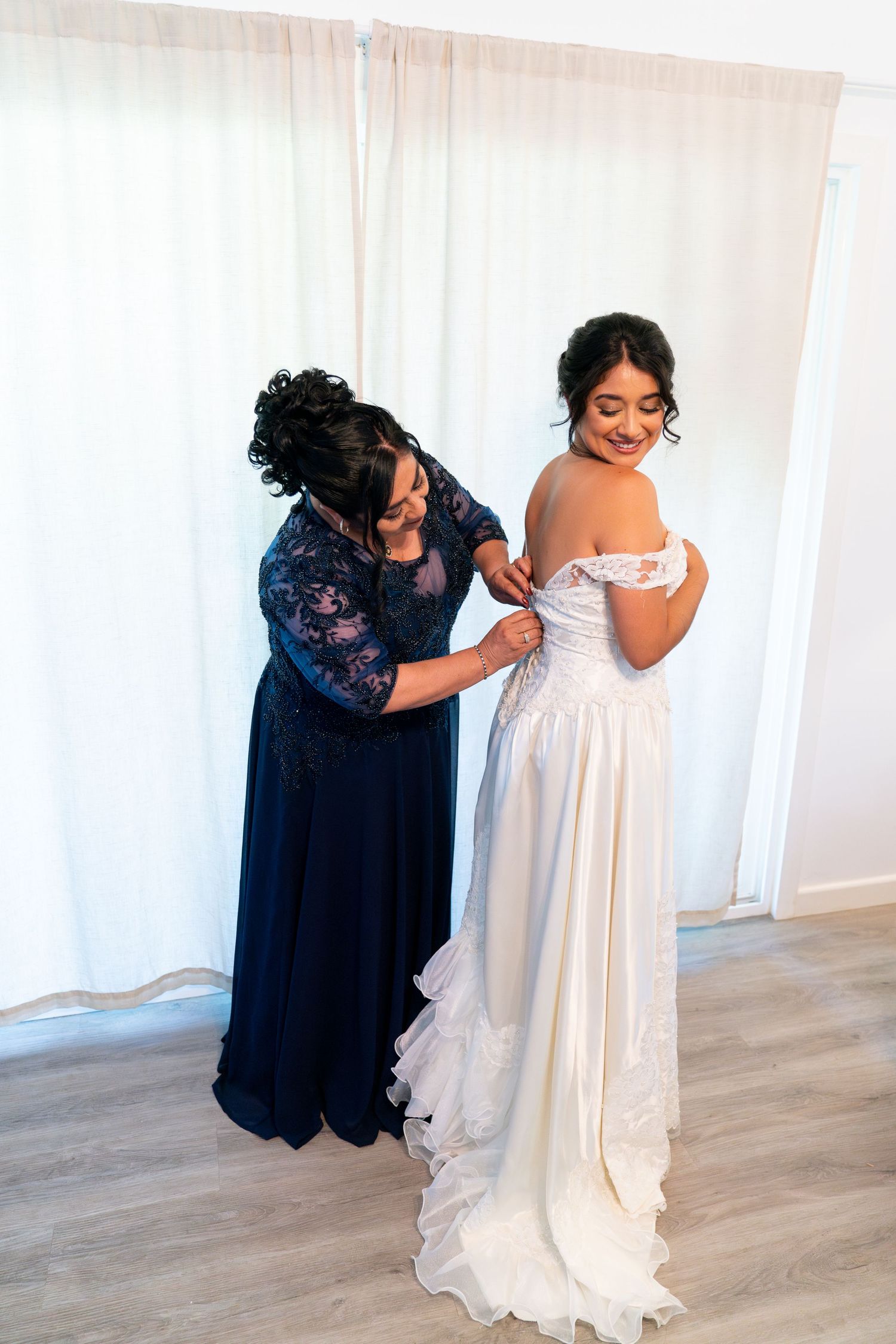 A woman in a navy blue dress helps adjust a white wedding gown on a bride in a brightly lit room.