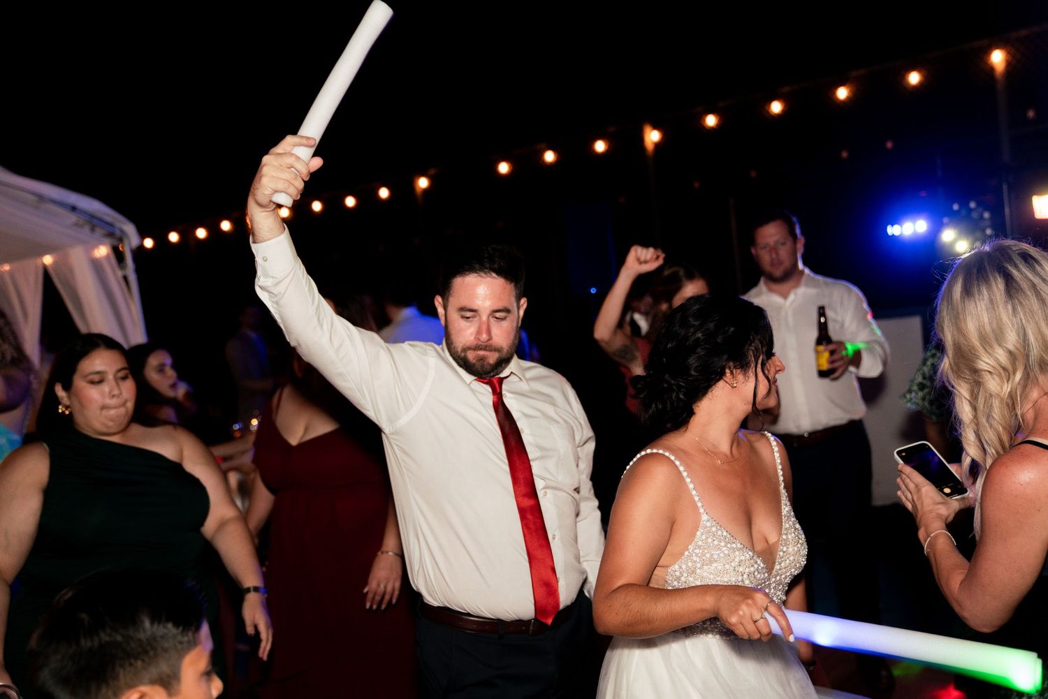 Wedding reception dance party with guests holding light sticks under string lights at night.
