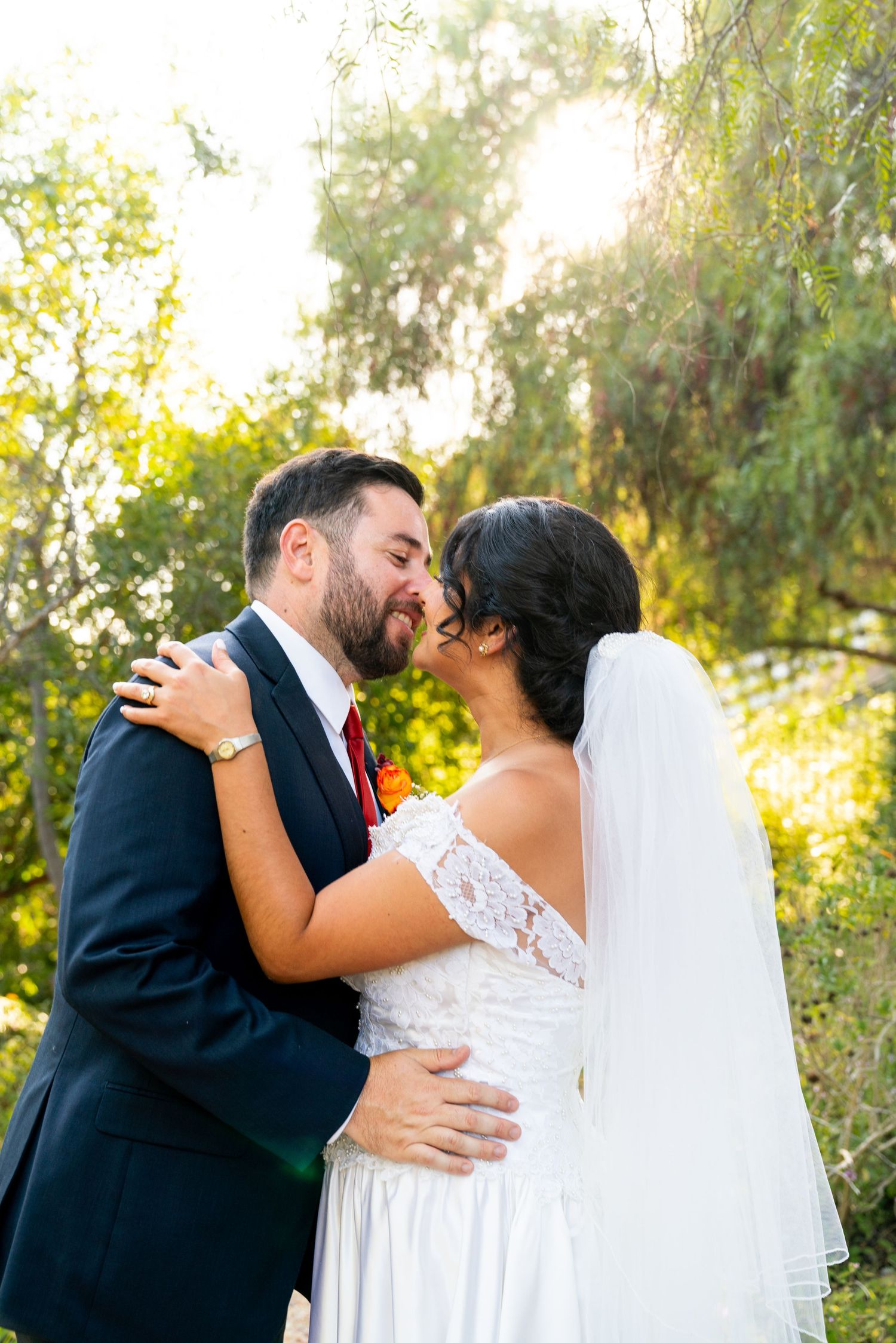 A newlywed couple share an intimate moment in a garden setting with golden sunlight filtering through trees.