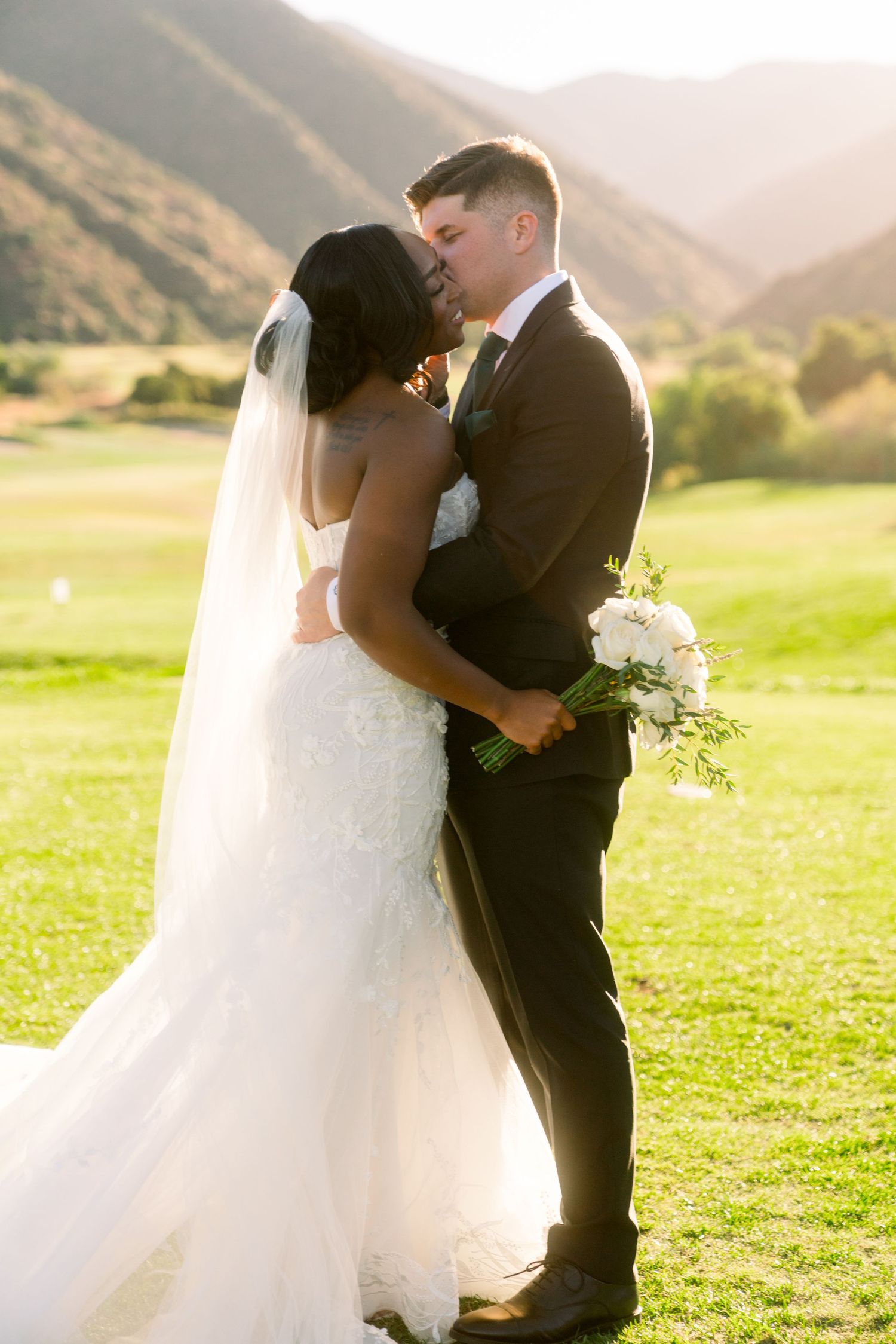 Romantic wedding couple embracing on grassy field with mountain landscape at sunset.