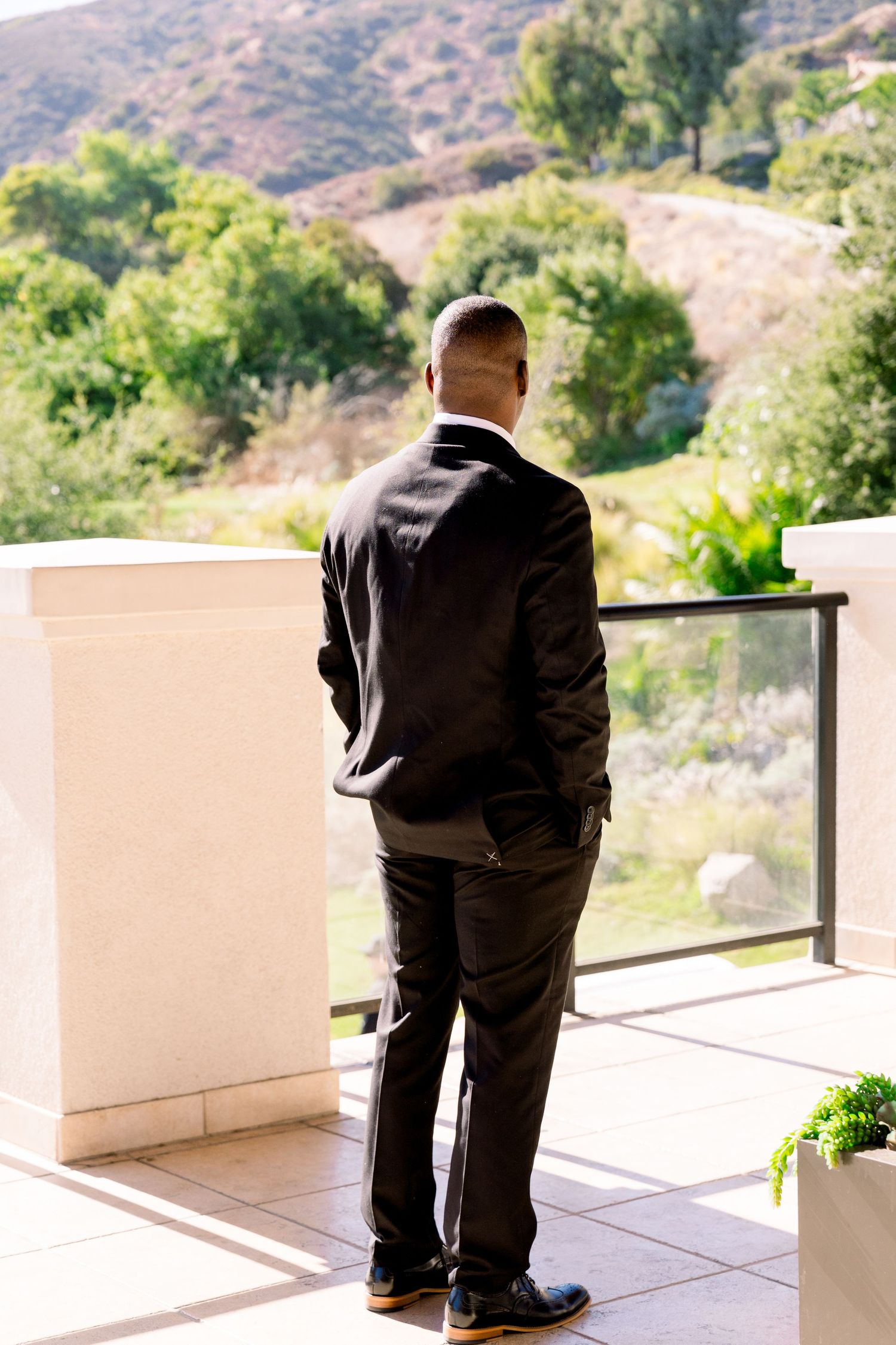 A person in a dark suit stands on a balcony overlooking mountainous terrain on a sunny day.