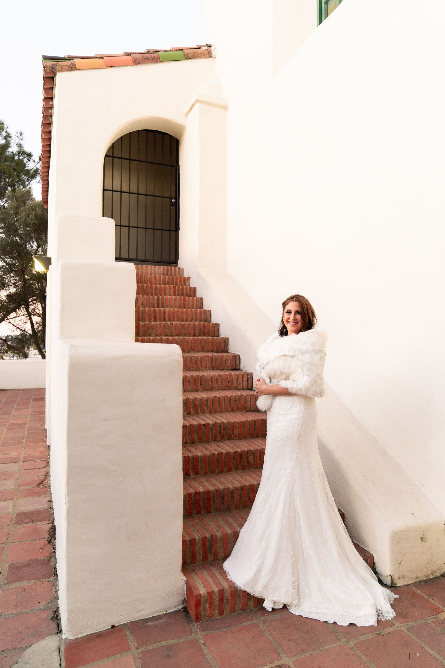 A bride in a long white wedding dress and fur wrap stands elegantly on brick steps against a cream stucco wall.