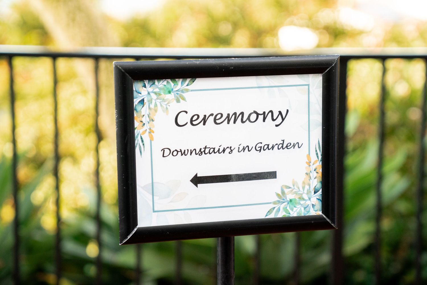 A framed directional sign for a garden ceremony features floral accents and black text against a white background.