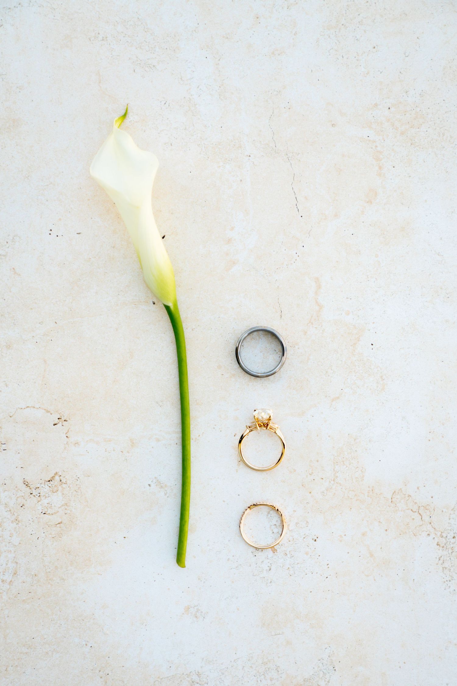 White calla lily flower arranged with two gold rings and one silver ring on a light textured background.