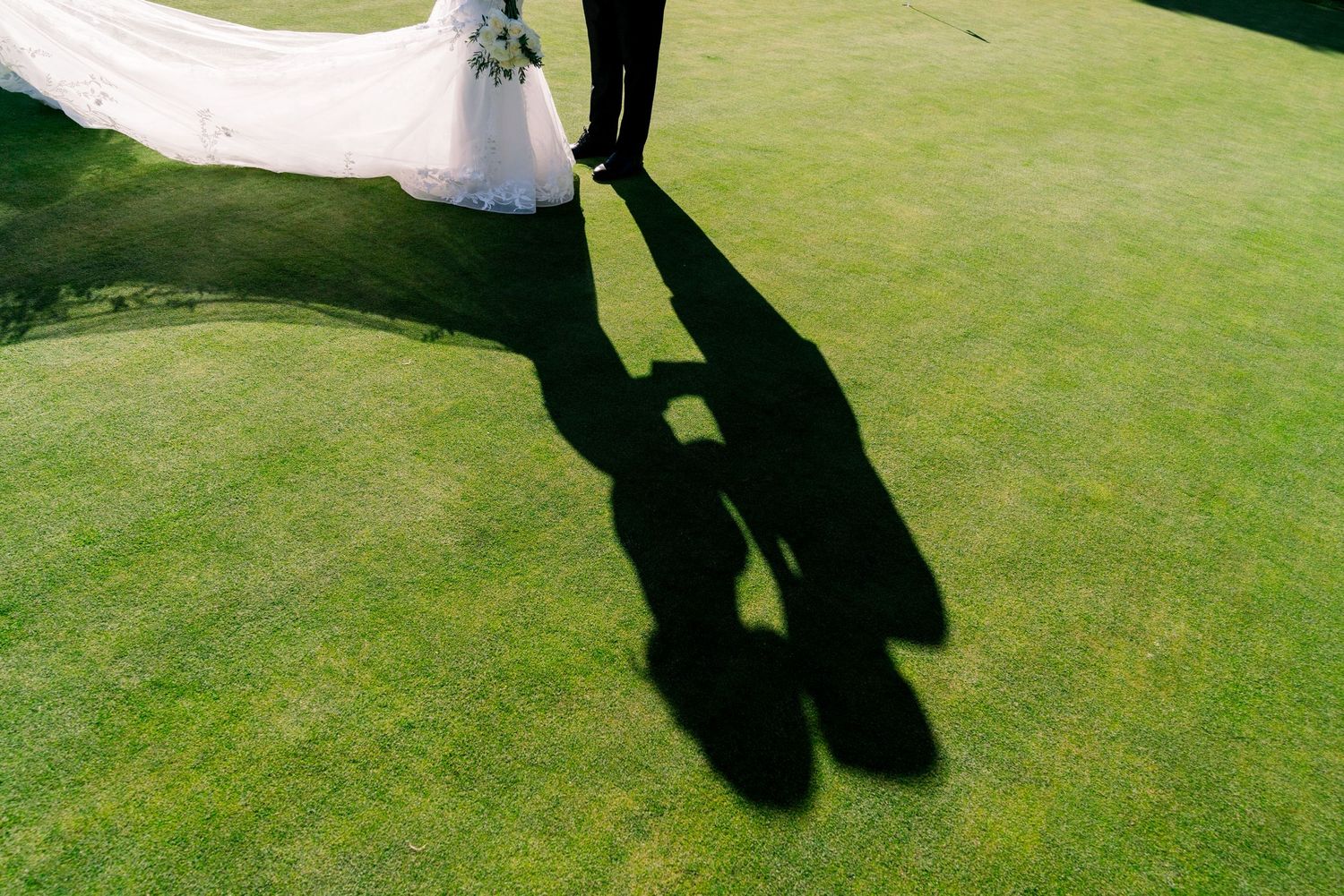 Shadows of a wedding couple cast on green grass with flowing white dress visible.