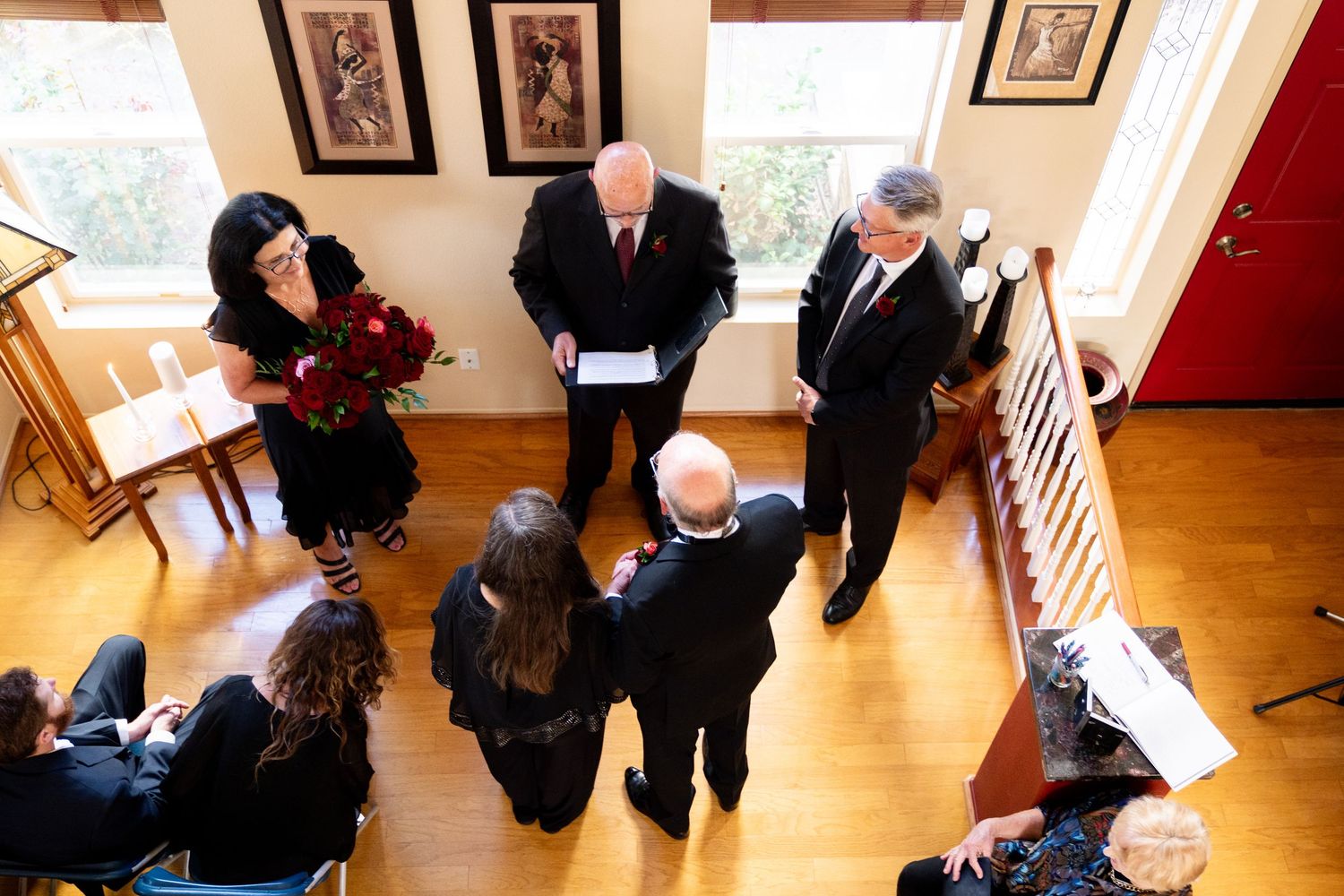 People gather in a bright indoor room with hardwood floors and framed artwork on the walls during a formal gathering.